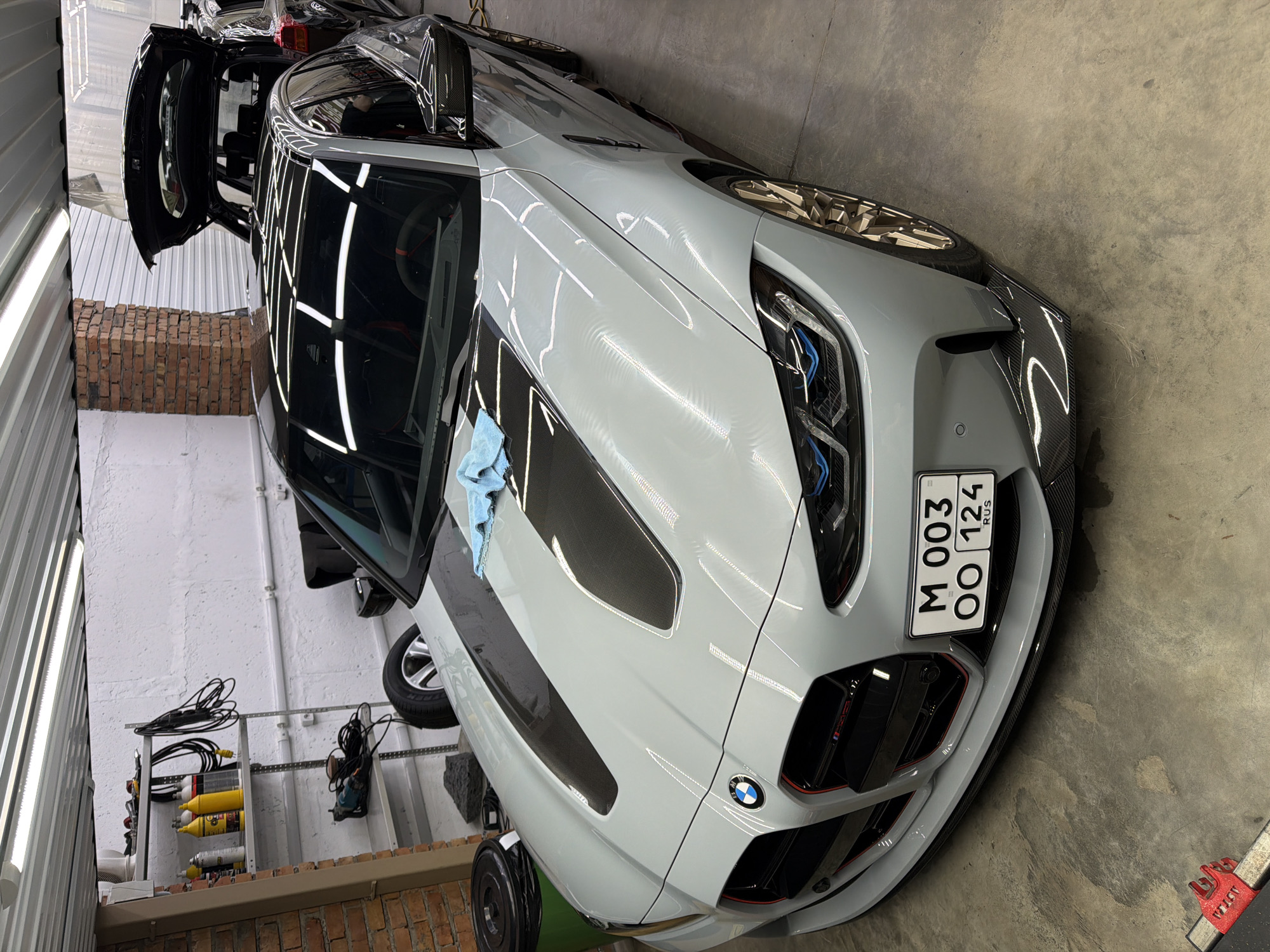 The image shows a light gray BMW with an open door parked in what appears to be a garage or workshop. The car has carbon fiber accents on the hood and front. A blue cloth sits on the hood. The license plate reads "M 003 OO 124 RUS". The environment includes a wall with shelves holding equipment, a concrete floor, and fluorescent lighting. The vehicle is likely located in Krasnoyarsk, Russia based on the license plate.