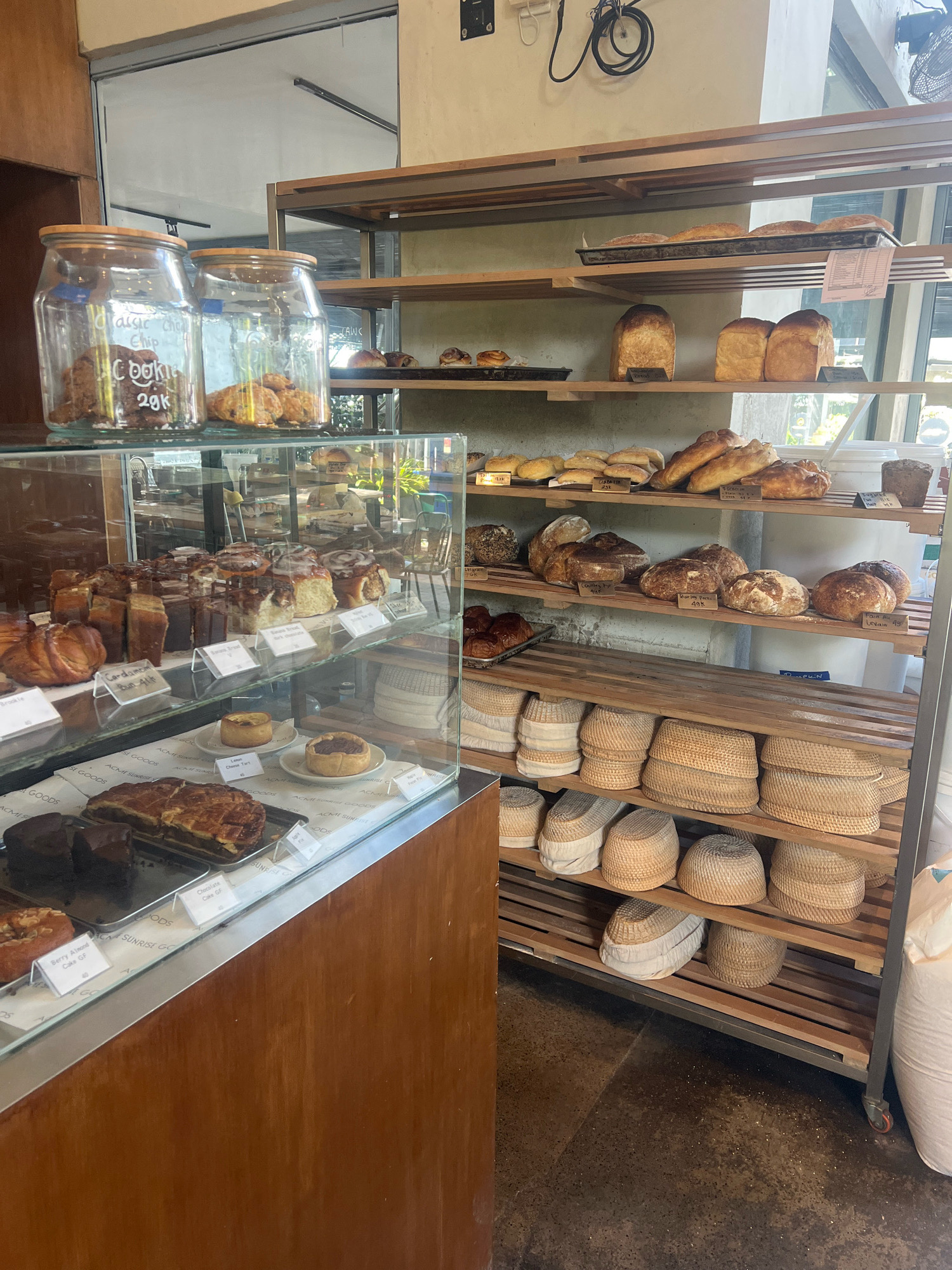 The image shows the interior of a bakery in North Kuta, Indonesia. The bakery features wooden shelves displaying a variety of baked goods, including loaves of bread, pastries, and cookies. There are also glass display cases filled with cakes, tarts, and cinnamon rolls. Several woven baskets are stacked on lower shelves. The overall impression is of a well-stocked and inviting bakery.