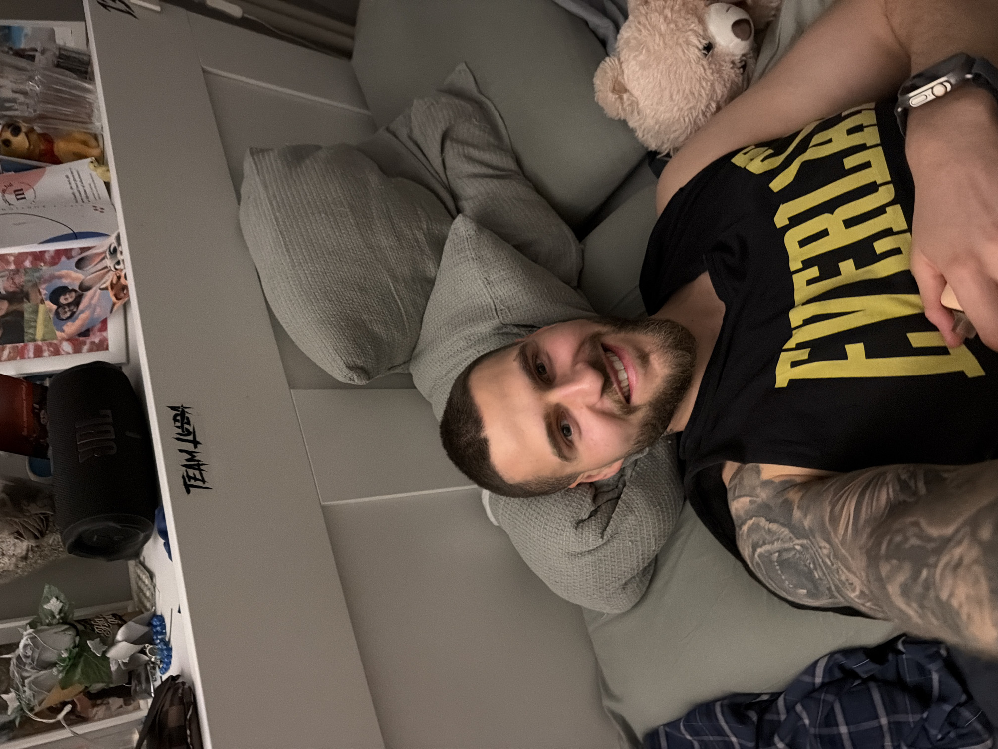 The image shows a smiling young man, likely in his 20s or 30s, relaxing indoors on a grey couch or bed. He has short dark hair and a beard and is looking directly at the camera with a broad, cheerful smile. He is wearing a black t-shirt with yellow text, partially visible as "FIGHTER." On his left wrist, he wears a silver Apple Watch with a dark band. His left arm is extensively covered with a detailed greyscale tattoo, appearing to depict a roaring animal, possibly a wolf.
He is surrounded by several grey pillows, some with a waffle-knit texture, and a light brown teddy bear rests near his head. To his left, a white shelving unit holds various items. The top shelf contains a yellow Winnie the Pooh plush toy and papers, one with an "M" logo. The middle shelf prominently displays a black JBL portable speaker with its logo visible. The bottom shelf holds miscellaneous items, including a small brown bag with a patterned strap. A blue and white plaid blanket is visible at the bottom right. On the white surface of the shelf, the stylized text "TEAM TUDA" is written. The setting is a casual indoor room, possibly in Sofia, Bulgaria.