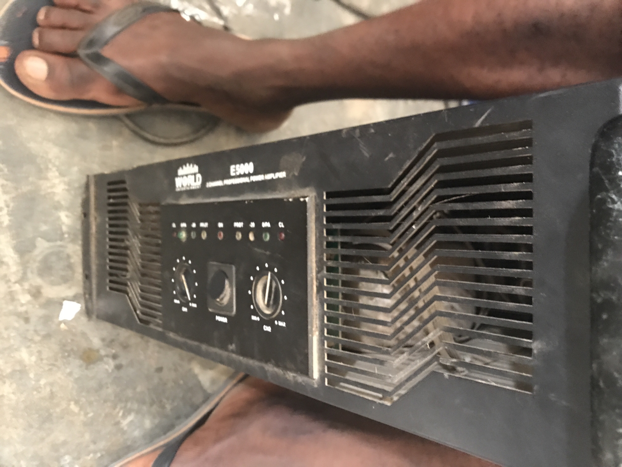 An African male with dark skin, wearing a black flip-flop on his right foot, is standing near a black professional power amplifier labeled "ES5000". The amplifier has a grated ventilation system on its upper section and a control panel on the lower section with knobs and indicator lights. The surrounding environment appears to be an indoor or covered outdoor space with a concrete floor. The image focuses on the lower portion of the male's body and the amplifier, suggesting the photo was taken at a close range. The location is identified as New Stadium, Nigeria.