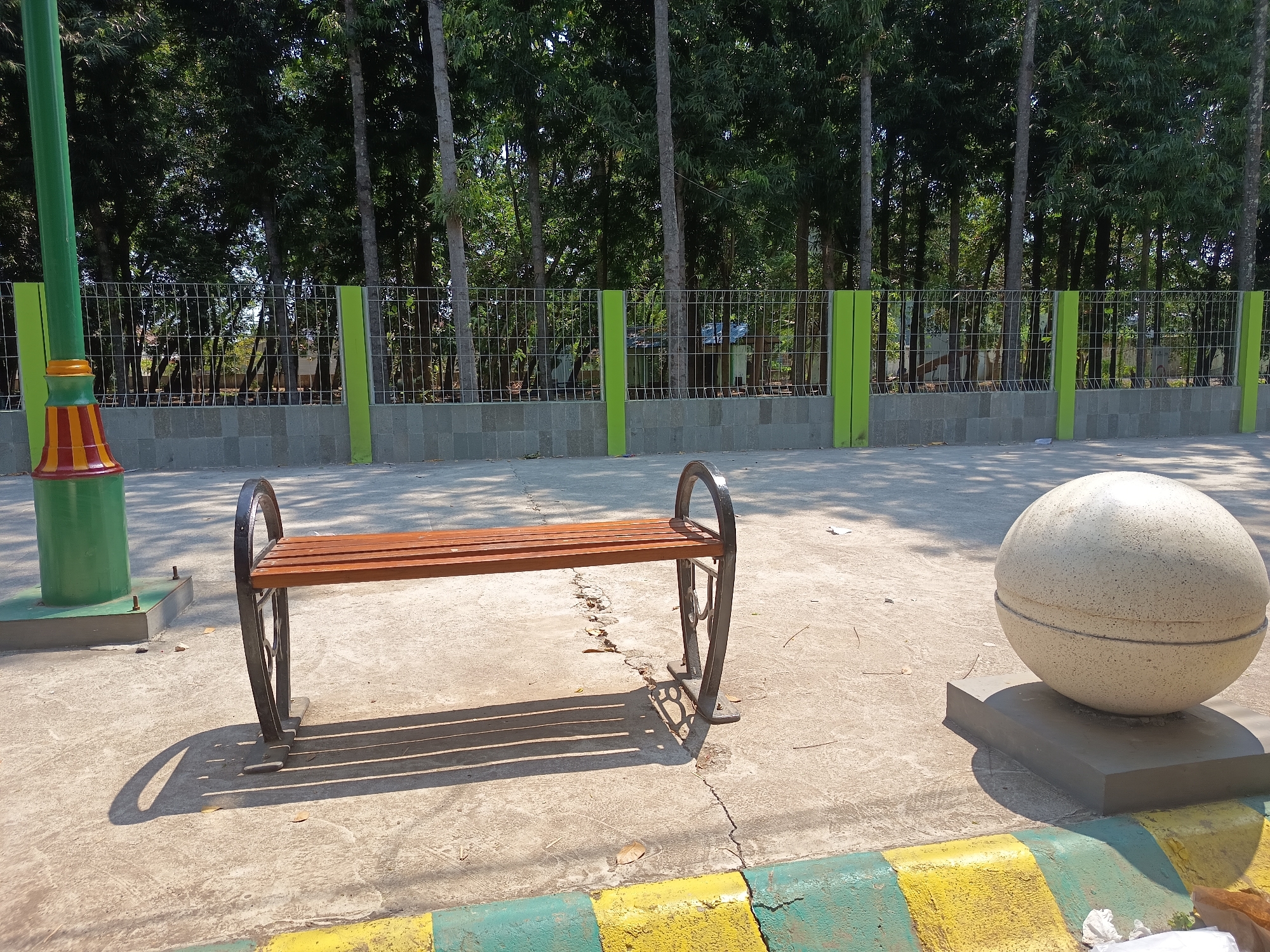 The image shows an outdoor scene with a park bench and a large spherical concrete object in the foreground. The bench is made of wood and metal, with curved metal legs. The spherical object is light-colored and rests on a dark gray base. Behind them, a metal fence with green posts separates the paved area from a line of trees. To the left, a green lamppost with colorful accents is visible. The ground is paved with concrete and cracked in places. The scene appears to be a public space, possibly a park or a garden. The location is identified as Kandangjati Kulon, Indonesia. There are no people or animals in the image, and no actions or interactions are taking place.