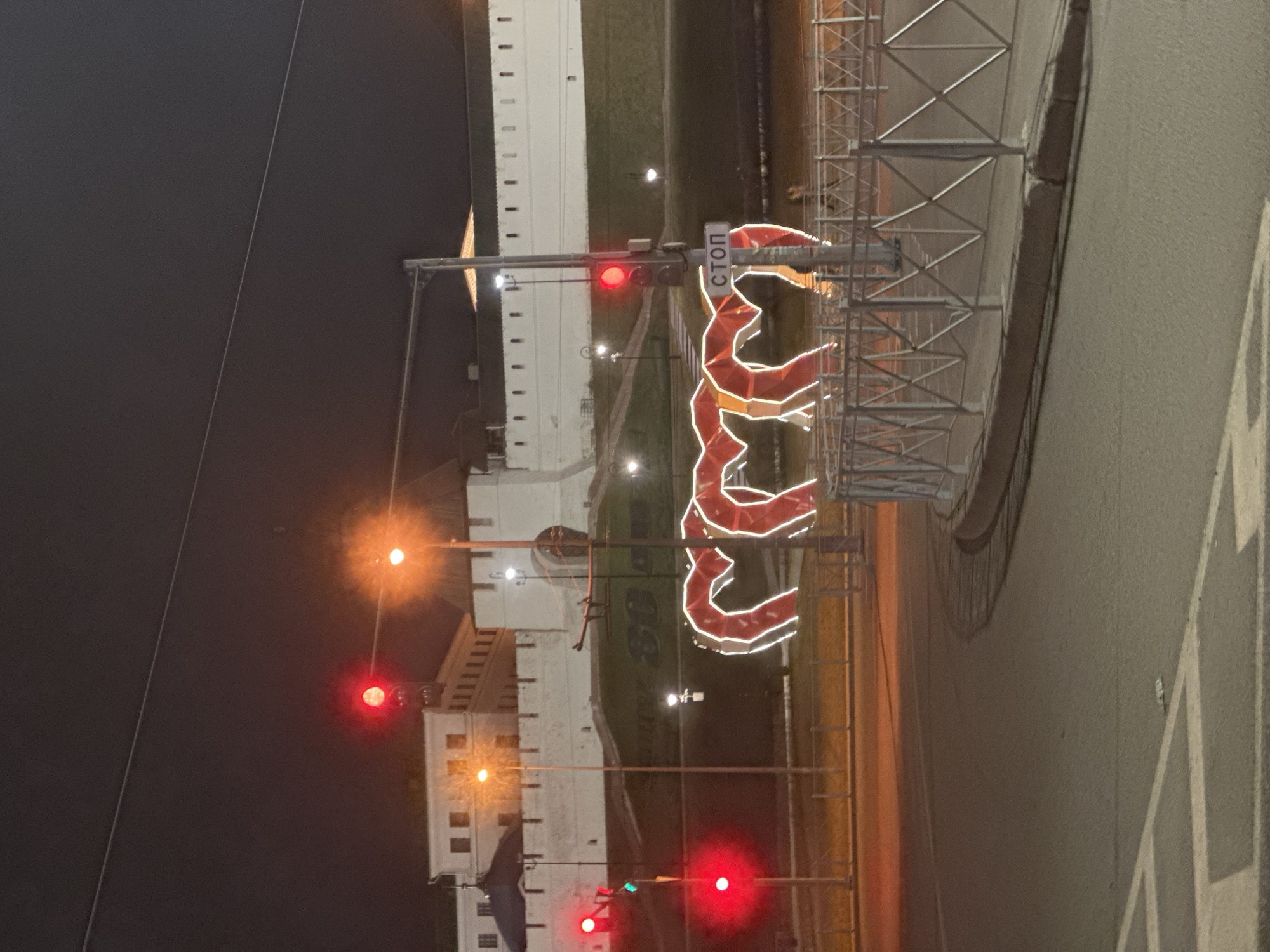 The image depicts a night scene in Kazan, Russia, showing a cityscape with illuminated structures and a traffic light system. In the background is a white-walled structure with multiple windows, seemingly a historic building or fortress. In front of the building, several traffic lights are visible, all showing red lights. There is also a sign in Russian that says "STOP". The image appears to be taken from ground level, capturing the intersection of a street with a walkway or entrance point to the building or fortress. The photograph is composed with a slightly off-center perspective.