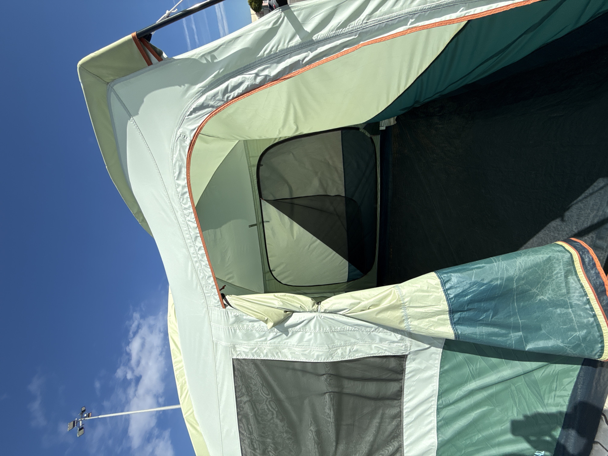 The image shows an open, light green tent set up outdoors. The tent has a mesh window on the left side and an open entrance leading into the interior, which also has a smaller zippered opening. The tent fabric has sections of white, light green, and dark green. The ground visible inside the tent is dark. In the background, a bright blue sky with scattered white clouds is visible. A tall pole with multiple lights on top is also present in the upper left quadrant of the image. The scene appears to be in a relatively open area, possibly a campground or a park. There is no indication of the city of Bielany Wrocławskie, Poland, in the image.