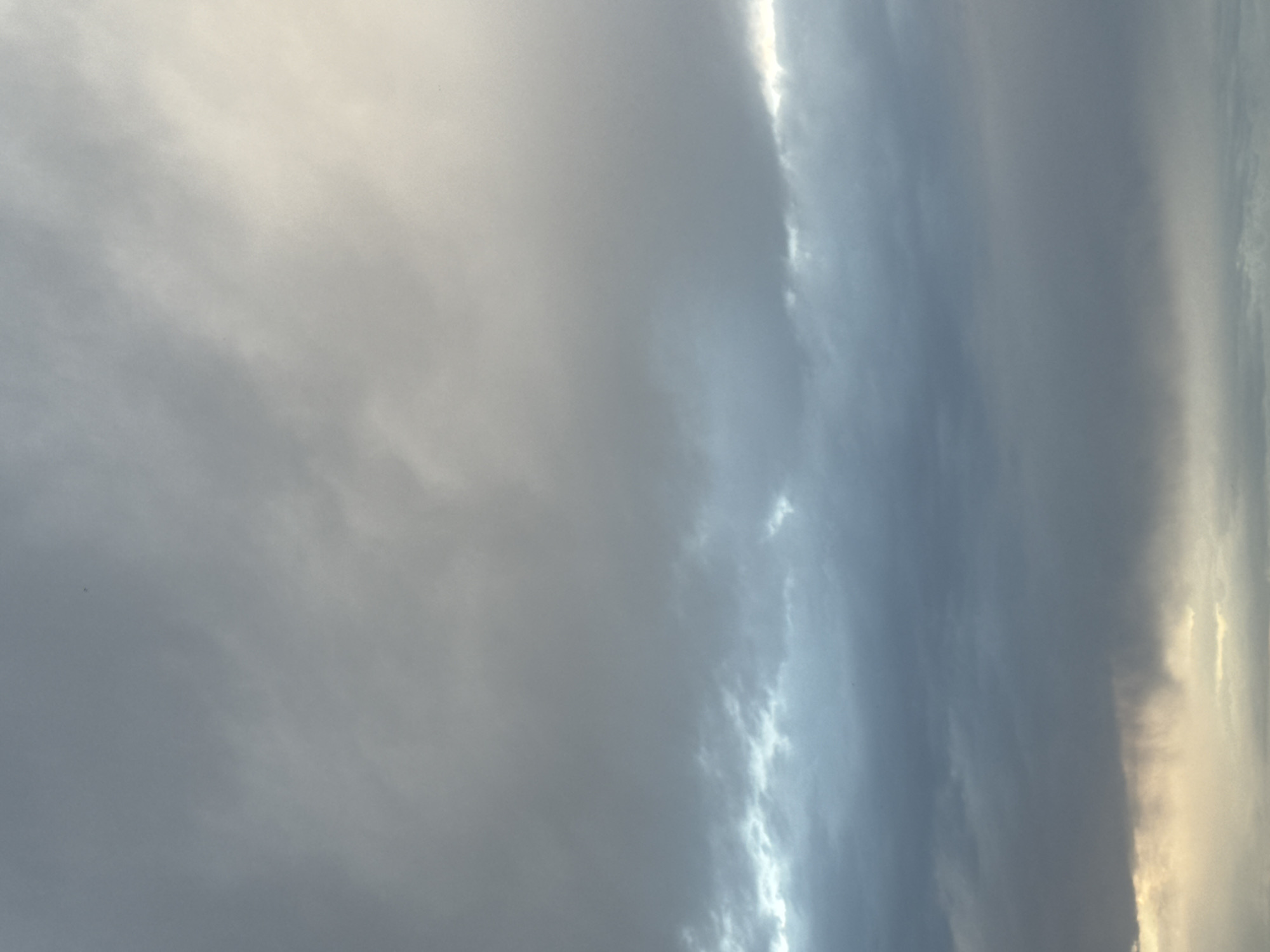 The image depicts a cloudy sky above Bishkek, Kyrgyzstan. The clouds are layered, with darker, heavier clouds in the lower half of the frame and lighter, more diffused clouds in the upper half. There are streaks of rain visible in the lower center of the image, indicating active precipitation. The lighting suggests it is either dawn or dusk, with a faint glow on the horizon. No people, specific structures, or identifiable landmarks are visible, making it a general atmospheric scene.