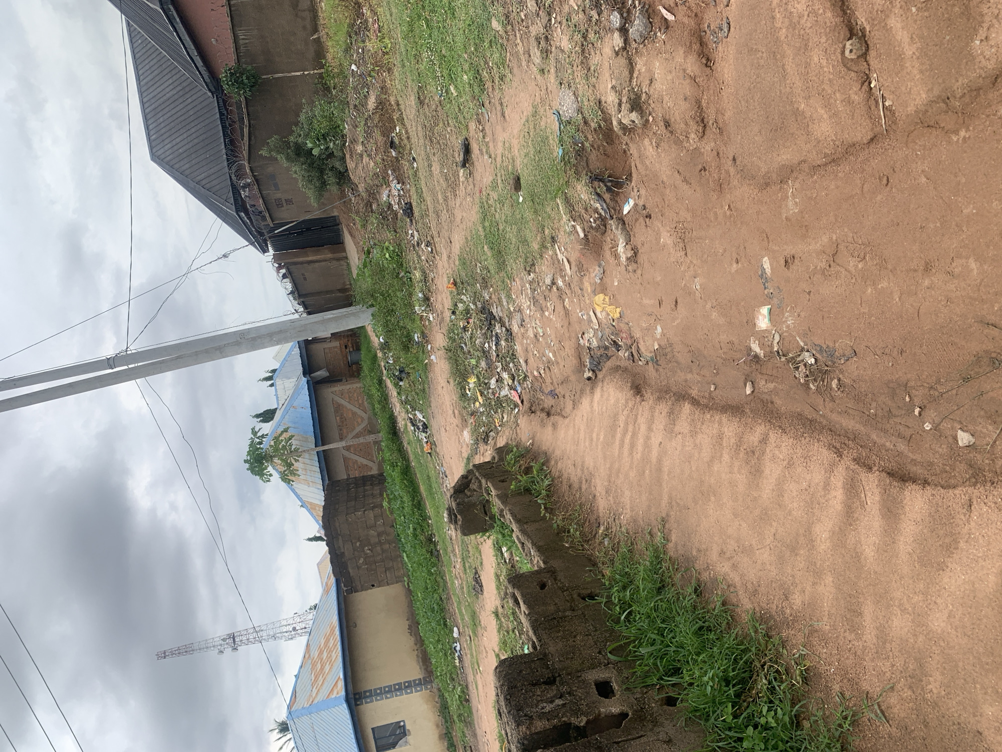 The image depicts a dusty, unpaved road in a residential area of Jalingo, Nigeria. The road is lined with houses, some with blue corrugated metal roofs and others with darker, sloped roofs. A prominent cellular tower is visible in the distance, indicating the presence of telecommunications infrastructure. The environment appears somewhat neglected, with scattered litter along the roadside and in the drainage ditches. Vegetation, including grass and small trees, grows sporadically along the edges of the road and around the buildings. A concrete structure, possibly a damaged wall or foundation, is present in the foreground. The sky is overcast with clouds. The scene conveys a sense of an ordinary, everyday street in a Nigerian city.