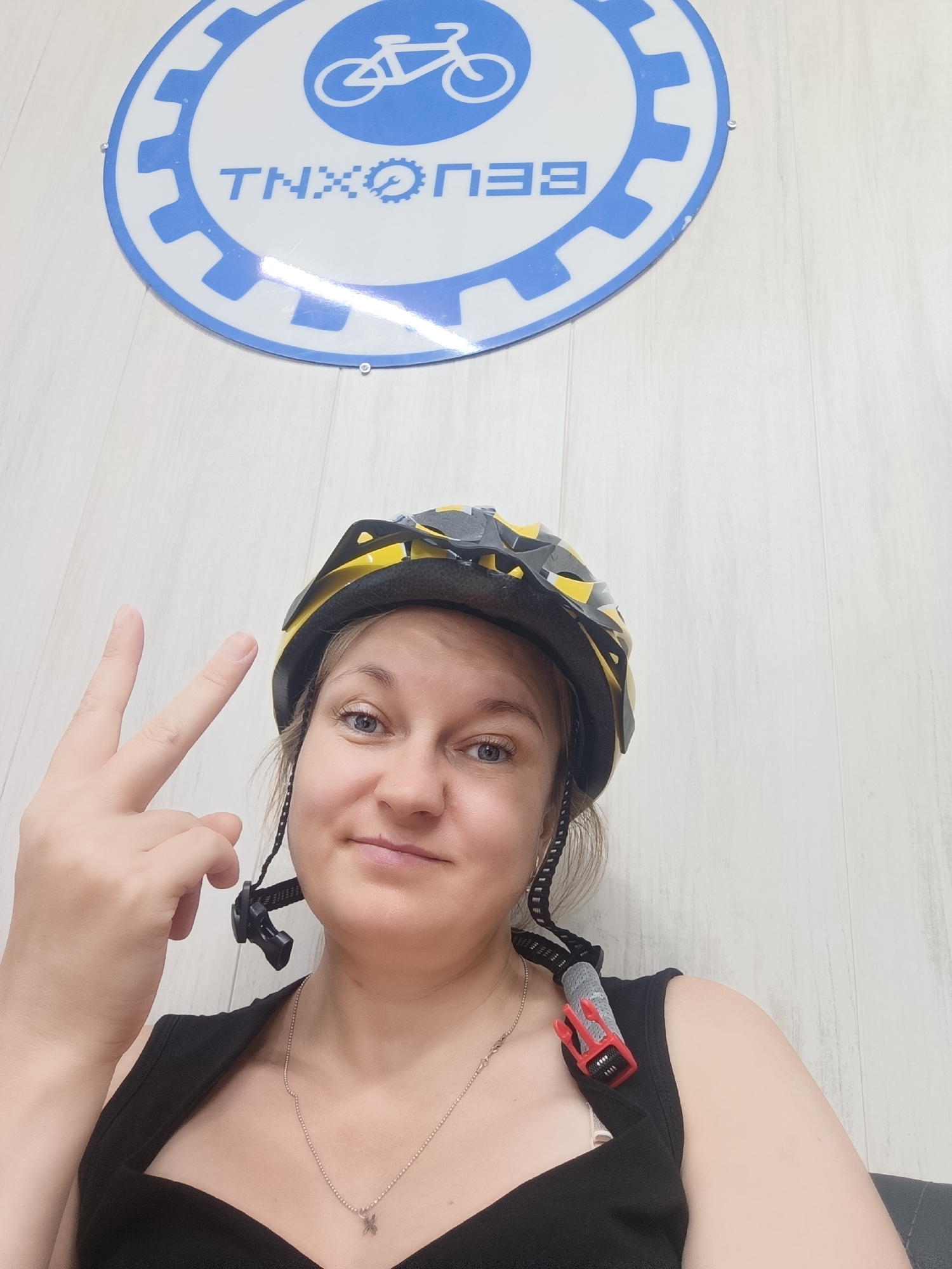 In this close-up, selfie-style photograph, a young woman with blonde hair and blue eyes is wearing a yellow and black bicycle helmet. She is looking at the camera with a slight smile and holding up two fingers in a peace sign. She is wearing a black tank top and a silver necklace with a small charm. The background is a white wooden wall with a blue and white circular logo featuring a bicycle and text that appears to read "TNH ORE3". The image was taken in Kostanay, Kazakhstan.