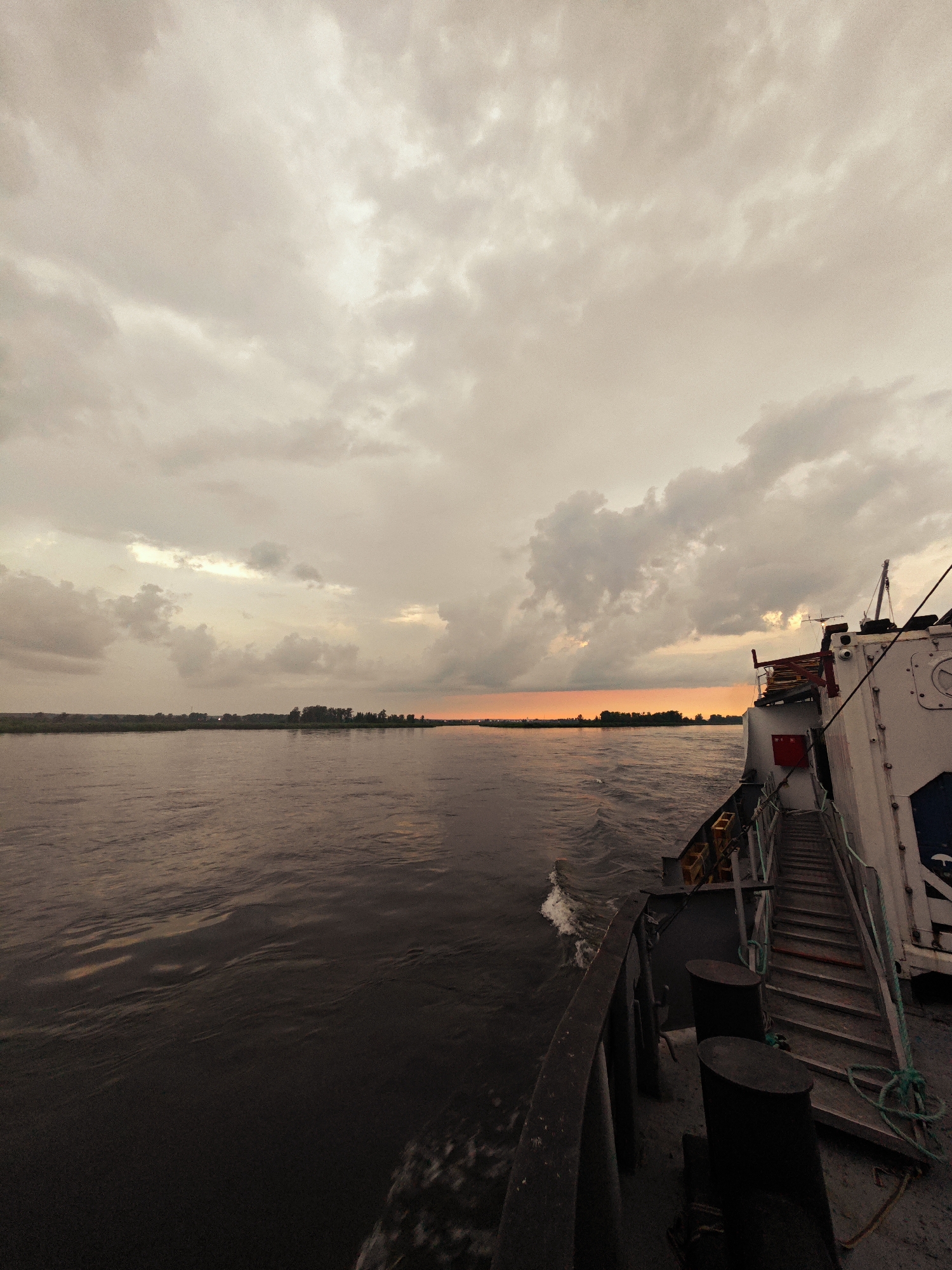A wide-angle, low-angle, vertical shot captures a river scene from the deck of a boat near Kazachinskoye, Russia. The sky is filled with overcast, grey clouds, with a sliver of orange light visible on the horizon, suggesting either sunrise or sunset. The river stretches across the frame, its surface rippling with dark, choppy water. On the far bank, a line of trees and vegetation forms a dark silhouette against the muted sky. The starboard side of a white vessel occupies the right portion of the image. A metal gangway with steps extends upwards along the side of the ship, and various industrial elements, including cleats and ropes, are visible on the deck. The boat appears to be in motion, with a small wake trailing behind it. The overall atmosphere is somber and overcast.