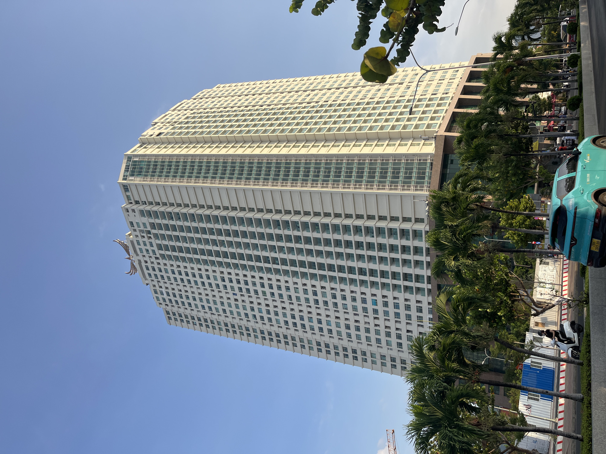 The image depicts a tall, multi-story building in Nha Trang Ward, Vietnam. The building is primarily white with many windows and has a modern architectural style. A decorative structure is visible near the top of the building. Palm trees line the street in front of the building, partially obscuring the lower levels. A blue car is parked on the street. Blue shipping containers and a parked motor scooter are also visible along the edge of the street. The sky is clear and blue.