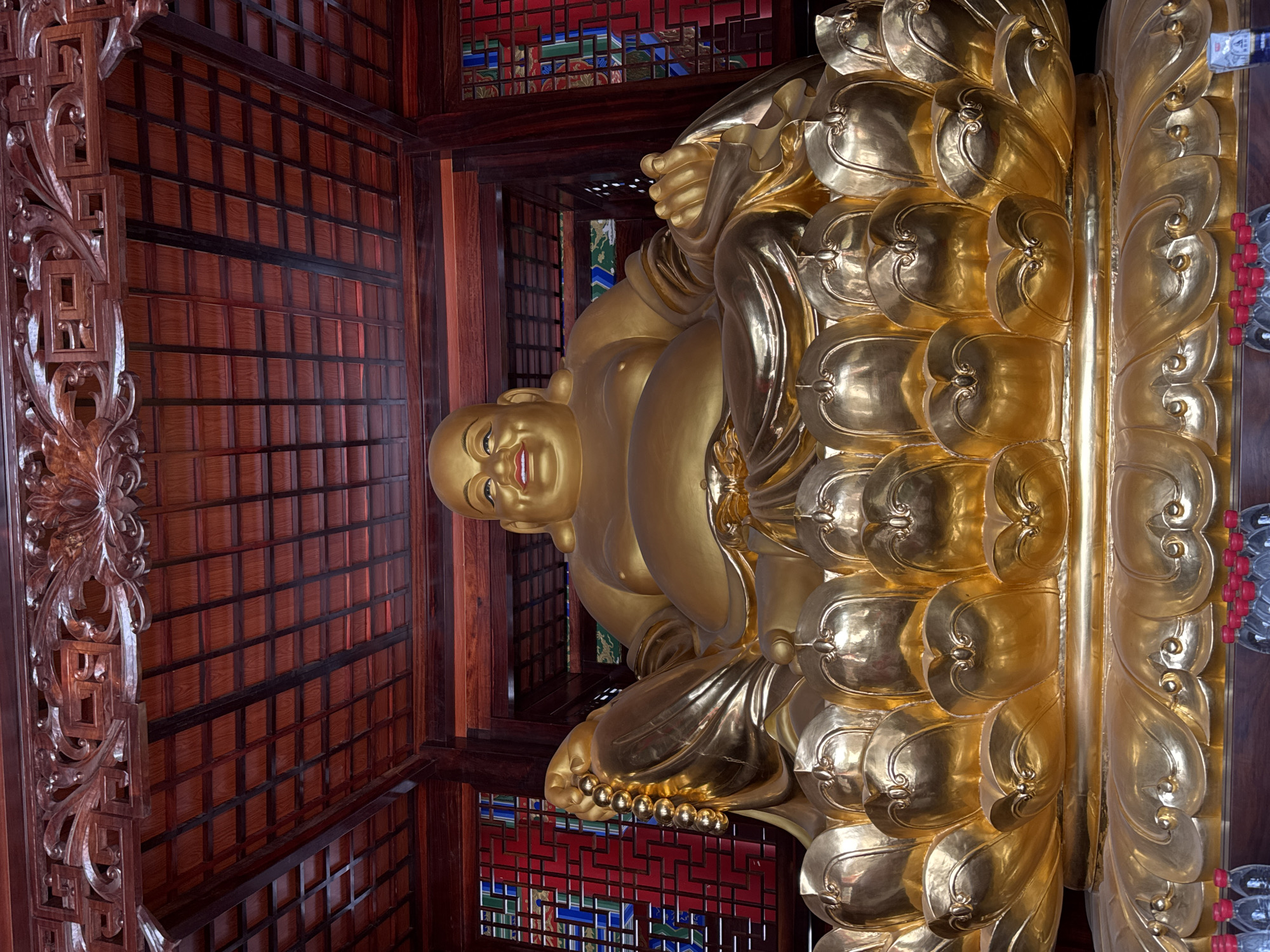 A golden statue of Buddha sits atop a lotus pedestal inside a room with a carved wooden ceiling and walls. The setting is likely a Buddhist temple in Qianjiang, China.