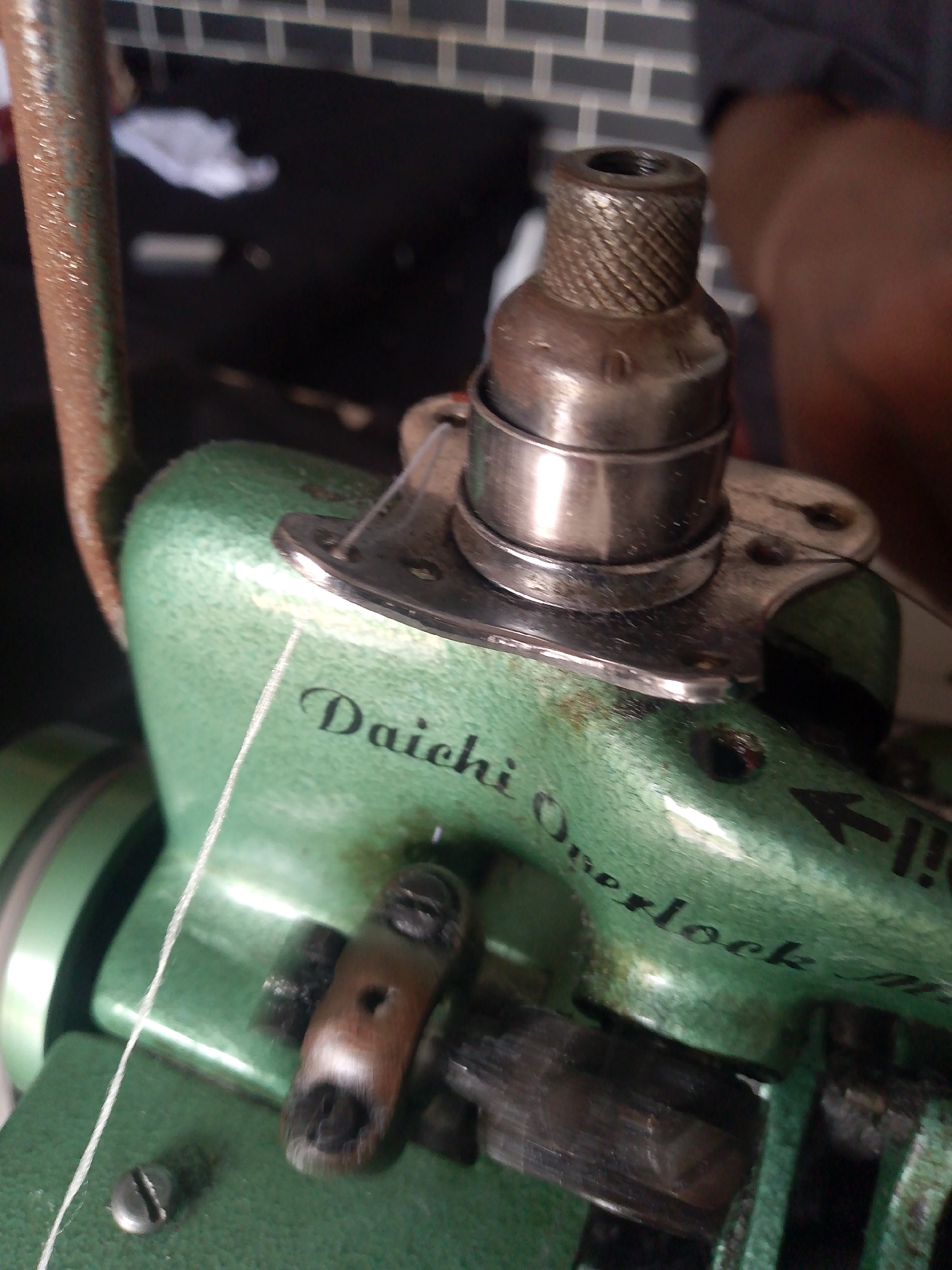 This is a close-up, low-angle, forensic photograph of a green sewing machine. The machine appears to be an older model, with some signs of wear and tear. A thin white thread is visible, running from the spool area down towards the machine's mechanisms. The words "Daichi Overlock" are clearly printed in black script on the green body of the machine. In the background, a blurry, dark area is visible, with what appears to be a brick-patterned wall and some lighter objects. No people are directly visible in the image, and no specific details of the location in Jauro Yinu, Nigeria, can be discerned from the visual information provided. The focus is on the mechanical components of the sewing machine, suggesting a potential need for repair or inspection.