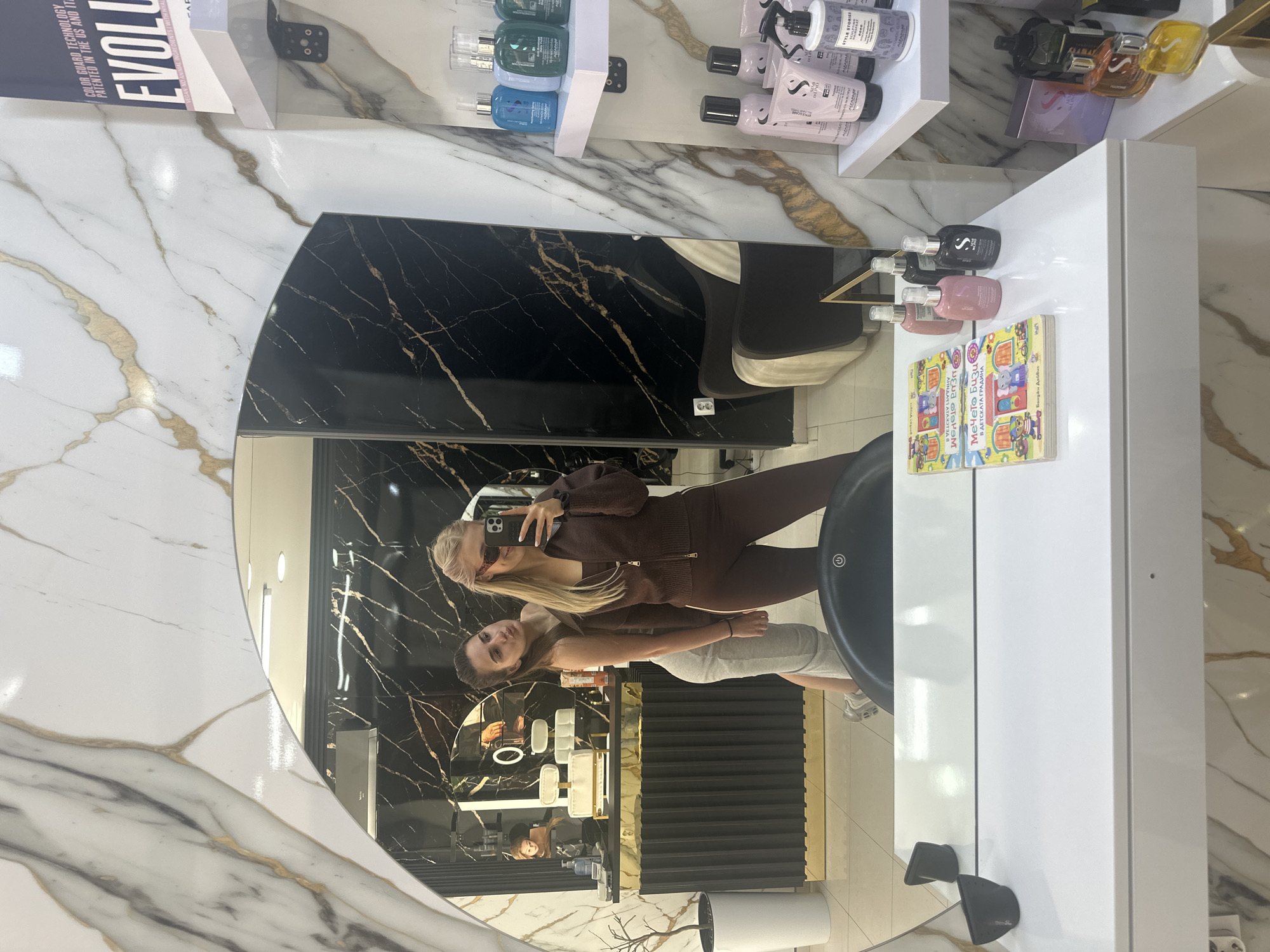 Here is a detailed description of the image:
**General Overview:** The image is an indoor shot, seemingly taken in a beauty salon or hair salon. The reflection in a large, round mirror dominates the scene.
**People:** Two women are visible in the mirror's reflection. One woman, with blonde hair, is holding a smartphone and appears to be taking the photo. She is wearing a brown tracksuit. The other woman is slightly behind her, with light brown hair, wearing a gray tank top and shorts.
**Objects:**
* **Mirror:** The focal point is a large, round mirror with a two-toned frame: white marble with gold veins and black marble with gold veins.
* **Salon Fixtures:** Behind the women are shelves with hair products, styling tools, and salon equipment. There's also a white cabinet next to the mirror.
* **Hair Salon Chair**: A hair salon chair with a black seat is visible.
* **Book**: On top of the white cabinet, next to the mirror, there's a children's book.
**Environment:** The location is a modern, stylish beauty salon with marble accents.
**OCR Text:** The OCR text reveals text from the products on the shelves and the title of the book, which suggests the location could be in Bulgaria, considering the book title is written in Bulgarian. Based on the book name "Мечето Бизи В ДЕТСКАТА ГРАДИНА", the language and local clues, it is likely in the city of Sofia, Bulgaria.