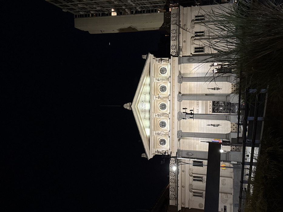 The "Escuela Presidente Roca" stands illuminated at night. Its classical facade features Doric columns and decorative medallions. The building's triangular pediment is prominently lit, contrasting with the dark night sky. A modern structure is visible atop the building, while the right side is partially obstructed by foliage. Street signs are also present.