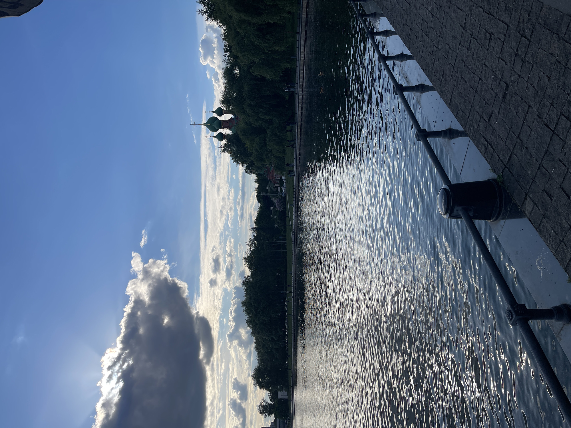 The image captures a waterfront scene in Unknown, Unknown. A body of water with rippling surface occupies the right half of the frame, bordered by a grey, tiled walkway with a black metal railing. Beyond the walkway, a line of trees is visible, silhouetted against a bright sky filled with scattered clouds. In the distance, three green domes, likely belonging to a church or cathedral, rise above the treeline. A large, shadowed cloud dominates the left side of the sky, with the sun casting rays through its edges.