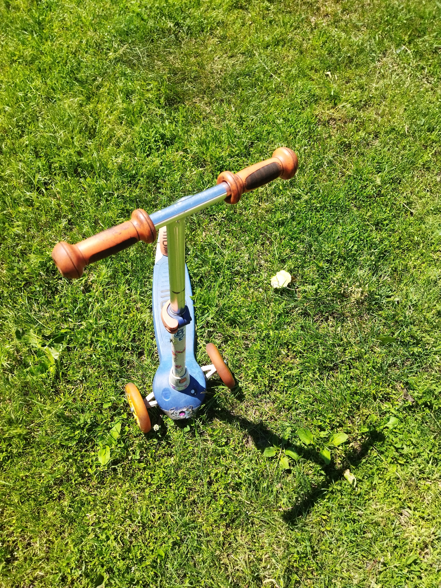 A blue three-wheeled scooter with brown handlebars is resting on a grassy area. The scooter has two orange wheels at the front and one at the back. The handlebars are silver with brown grips. The scooter appears to be unoccupied and stationary. The environment is an outdoor setting with abundant green grass, likely in a park or yard. Sunlight casts a shadow from the scooter onto the grass. There is a small light-colored object, possibly a fallen leaf or flower petal, in the grass to the right of the scooter. The scene is observed from a slightly elevated, downward-angled perspective.