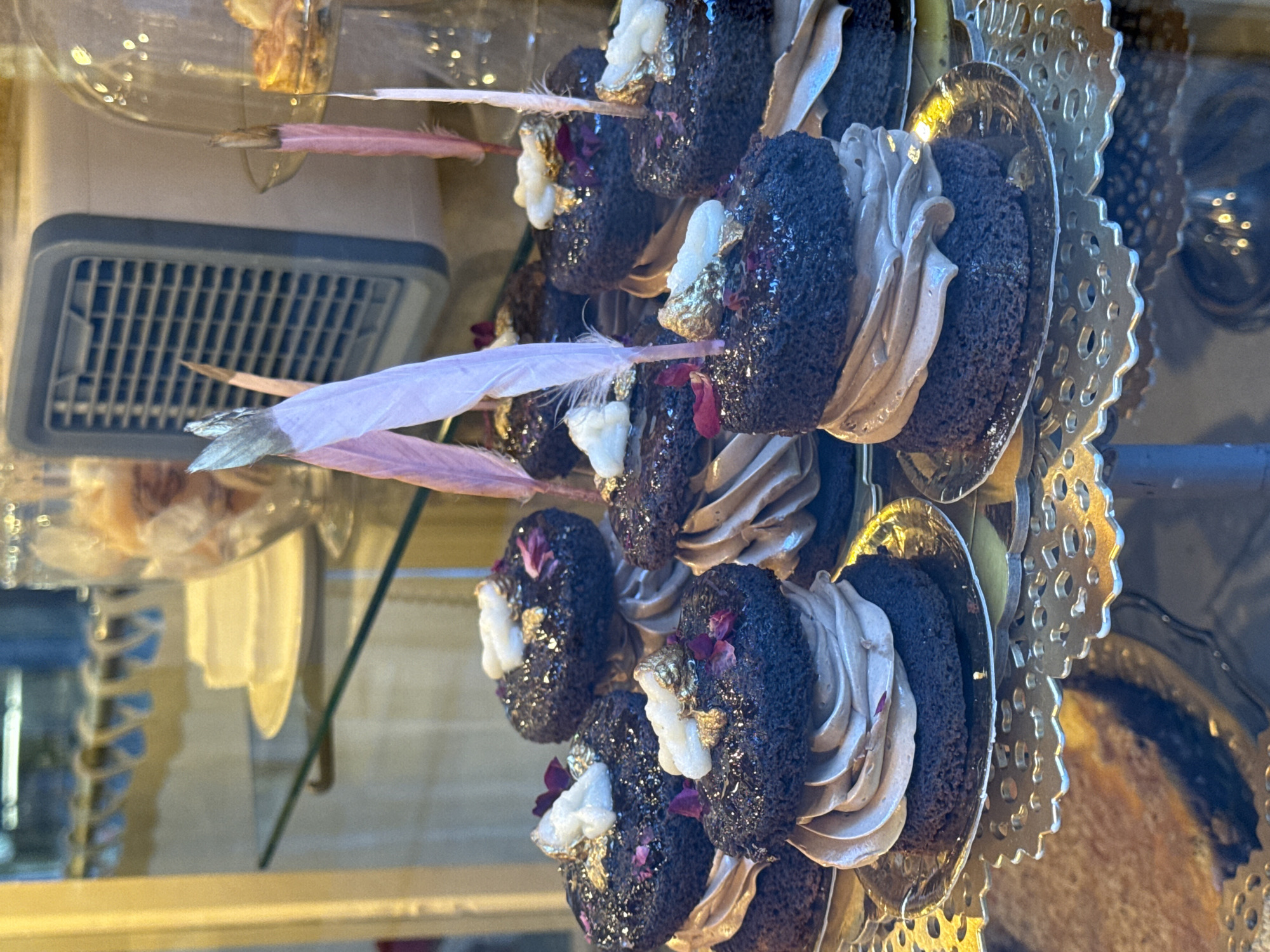 The image shows several rows of intricately decorated chocolate pastries inside a display case. The pastries are adorned with light brown frosting, edible gold flakes, and small pink feathers. They sit on individual ornate gold plates. The display case is likely located in a bakery or confectionery shop, and its ventilation system is visible in the background. Although the precise location in Barcelona cannot be determined from the visual data alone, these types of pastries are common in upscale bakeries in tourist areas of the city.