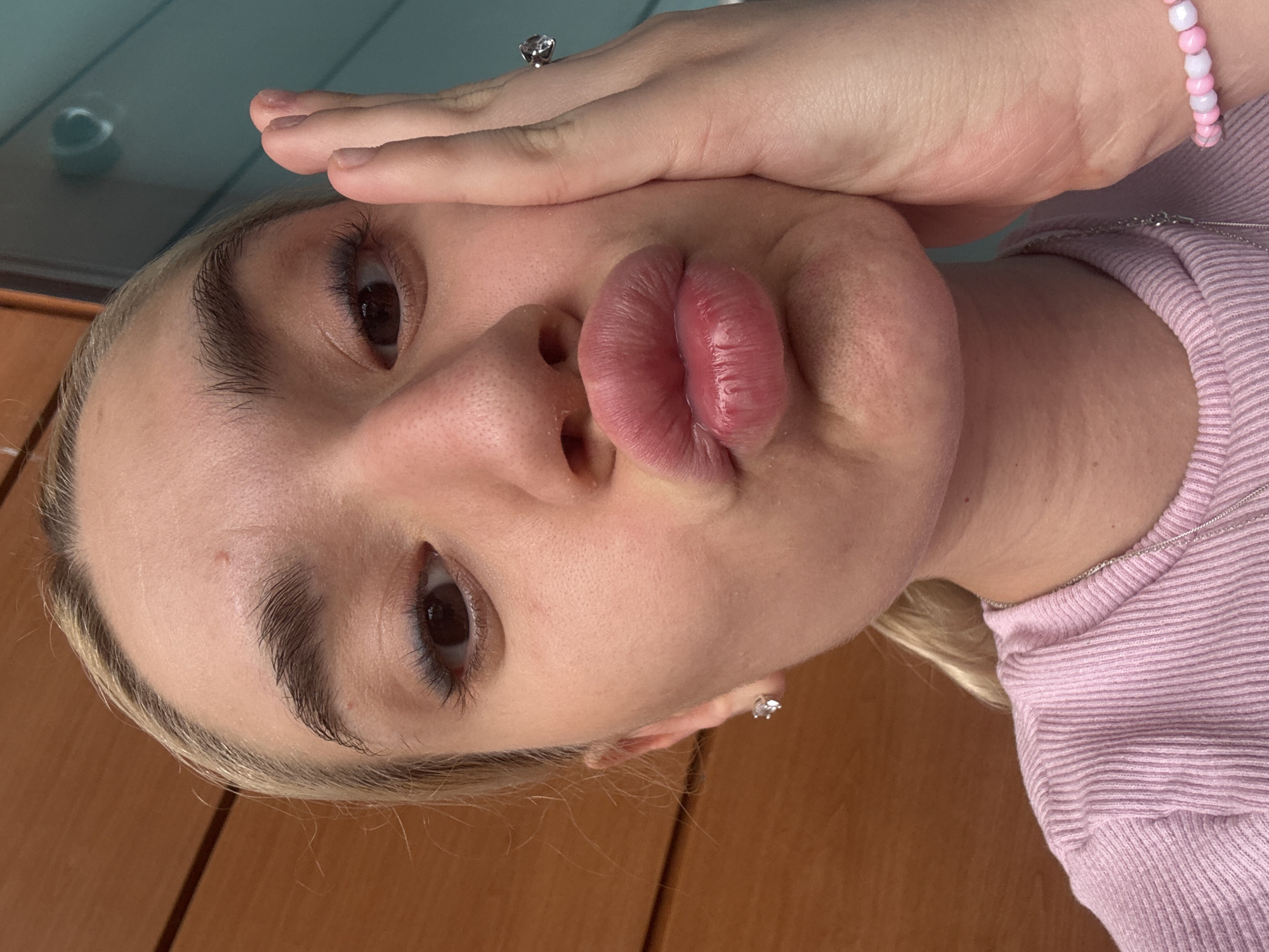 A close-up, rotated portrait shot features a young individual with fair skin and blonde hair, capturing a playful expression. The person's head is tilted significantly, with their face occupying a large portion of the frame. One eye, with a dark iris and long eyelashes, is clearly visible, while the other is partially obscured by a hand. The lips are prominently puckered into a "duck face" or kissy face, appearing full and pinkish-red. Dark, well-defined eyebrows frame the visible eye.
A hand, possibly the right hand, is placed across the forehead, covering part of the upper face. On one finger of this hand, a silver-colored ring with a clear, sparkling stone is visible. Another hand, on the right side of the frame, wears a pink and white beaded bracelet. A thin, silver-colored chain necklace rests against the skin, leading to a light pink, ribbed long-sleeved top. A small, clear stone stud earring is visible in one ear.
The background is simple, suggesting an indoor setting in Sofia, Bulgaria. The lower and right portions of the image show a light brown, possibly wooden, surface. The upper left corner reveals a muted teal or blue-green surface, possibly a wall or window. The lighting appears soft and natural, contributing to a candid, selfie-like aesthetic. The overall mood is lighthearted and casual, typical of a personal photo or social media post. No visible text is present.