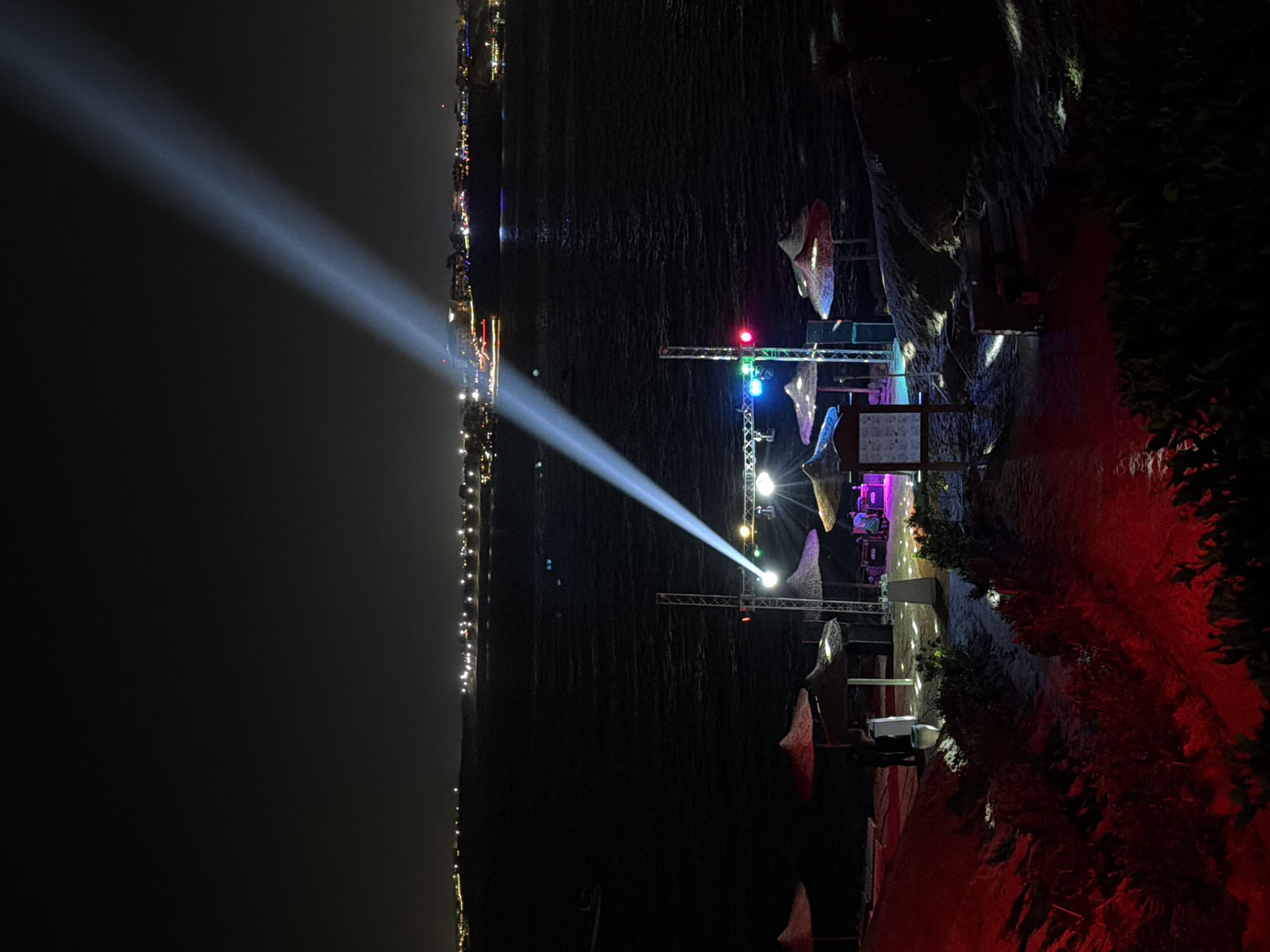 The image shows a nighttime view in Sharm El Sheikh, Egypt. On the left, the city skyline is visible with numerous bright lights lining the coast. A strong, white beam of light shines across the water from a structure on the right side of the frame. This structure appears to be a stage or performance area with lighting and possible decorative elements. Some people are visible near the base of the structure. The surrounding area on the right is dark, with hints of vegetation and perhaps a beach setting. Red lighting illuminates the ground at the base of the image. The water in between reflects some of the city lights and the beam.
