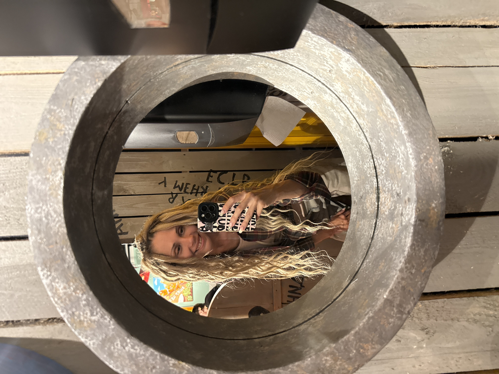 The image displays a woman taking a selfie, reflected in a large, circular, dark-grey metallic ring or mirror. The image itself is rotated 90 degrees clockwise, so the woman appears on her side.
The subject is a smiling woman with long, wavy blonde hair that flows across her chest. She looks directly at the camera with a cheerful expression. She is wearing a plaid shirt over a lighter top. In her right hand, she holds a black smartphone with a distinctive case featuring a pattern of repeating white or pink squiggly letters on a black background.
The circular frame is made of a textured, aged metal, possibly iron or steel, suggesting an industrial or rustic setting. Behind the woman's reflection, the background is composed of light-colored wooden planks or paneling. Visible text on these planks, reflected and inverted, includes fragments like "W E H y" and "E C L D". Other details reflected in the background include what appears to be a yellow surface, a white paper towel, and a colorful, blurry image, possibly a poster or artwork. A partial reflection of another person is faintly visible at the bottom of the circular frame.
The activity is clearly a self-portrait (selfie). The setting appears to be indoors with artificial lighting. The location context is Заволжье, Russia.