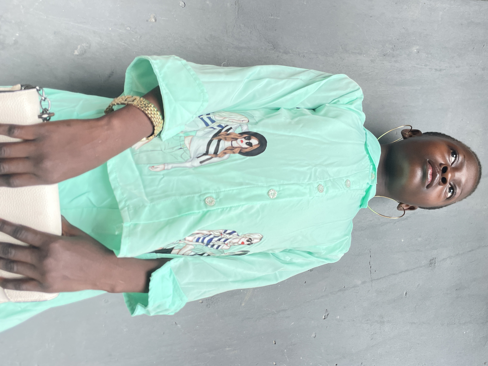 A light-skinned black female with short hair, wearing large hoop earrings, a light green button-down shirt with fashion illustration graphics, and a gold watch, is holding a light-colored bag with a chain. The picture is taken indoors on a grey-colored surface. The location is specified as Badagry, Nigeria.