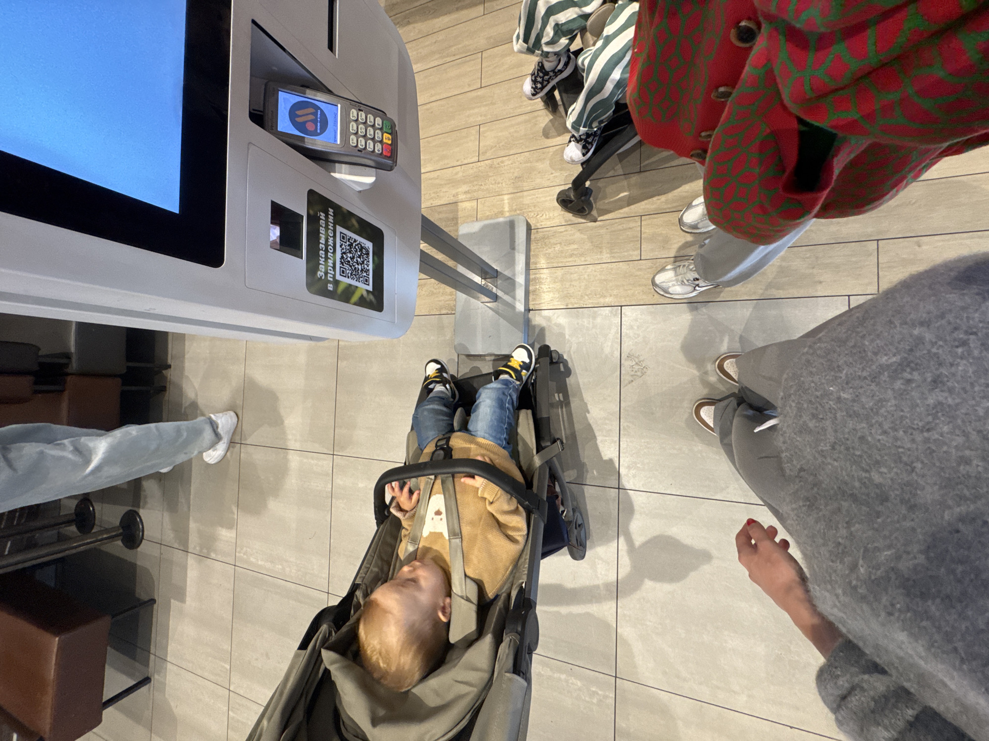 The image captures an overhead view inside a modern public establishment, likely a fast-food restaurant or a food court in Moscow, Russia. The setting features a light-colored tiled floor and contemporary furnishing elements.
In the foreground, a young child with blonde hair is seen comfortably reclined in a grey stroller, appearing to be asleep or resting. The child is dressed in a tan sweater with a white animal design, blue jeans, and black and yellow sneakers.
Surrounding the stroller are the legs and feet of several adults, indicating multiple people are present. One individual wears grey sweatpants and white sneakers, accompanied by a vibrant red and green patterned jacket or cardigan. Another person's legs are clad in light blue jeans and white sneakers. A third person wears grey attire with white and brown sneakers, and a hand with red nail polish is visible. The feet of a fourth person wearing green and white striped pants and black and white patterned sneakers are also seen, possibly from another stroller or chair.
To the left, a large self-service ordering kiosk dominates the space. It features a bright blue screen and an attached payment terminal displaying a logo and the Russian text "ВВОД-ОТМЕНА" (ENTER-CANCEL). Below the screen, a sticker with a QR code and the Russian phrase "Заказывай в приложении" (Order in the app) is visible.
The scene suggests a casual outing, possibly a family visit, with the child resting while the adults interact with the self-service facilities. The bright indoor lighting indicates daytime.
