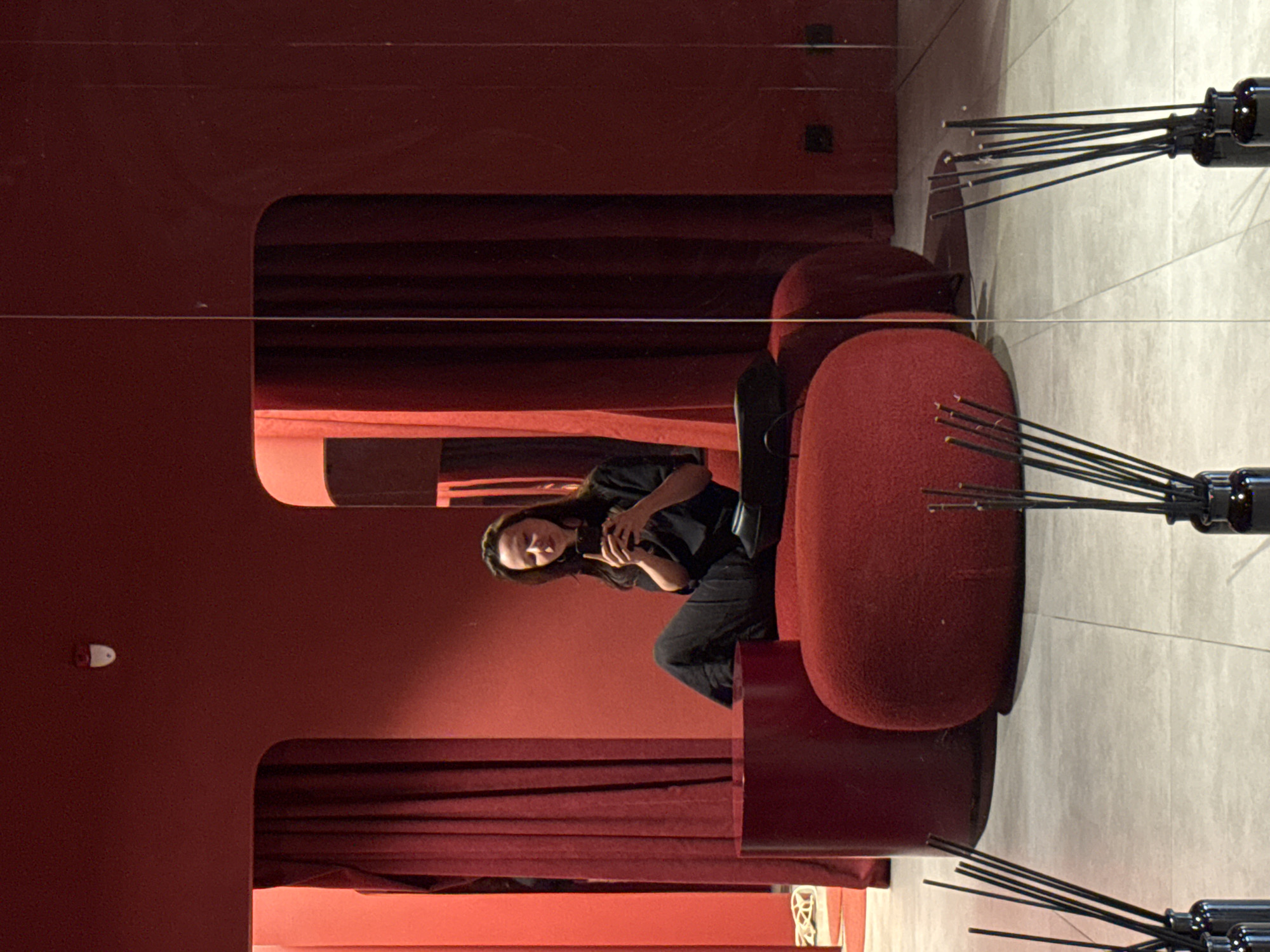 A woman is sitting on a red upholstered bench in a red-colored room, looking at her phone. She is wearing black clothing. In front of her is a red cylindrical table. To her right is another red upholstered object. The room has dark red curtains on the left and right sides. The floor is tiled in a light gray color. In the foreground, there are several bottles with reeds, suggesting they are reed diffusers. The image appears to be taken in Moscow, Russia.