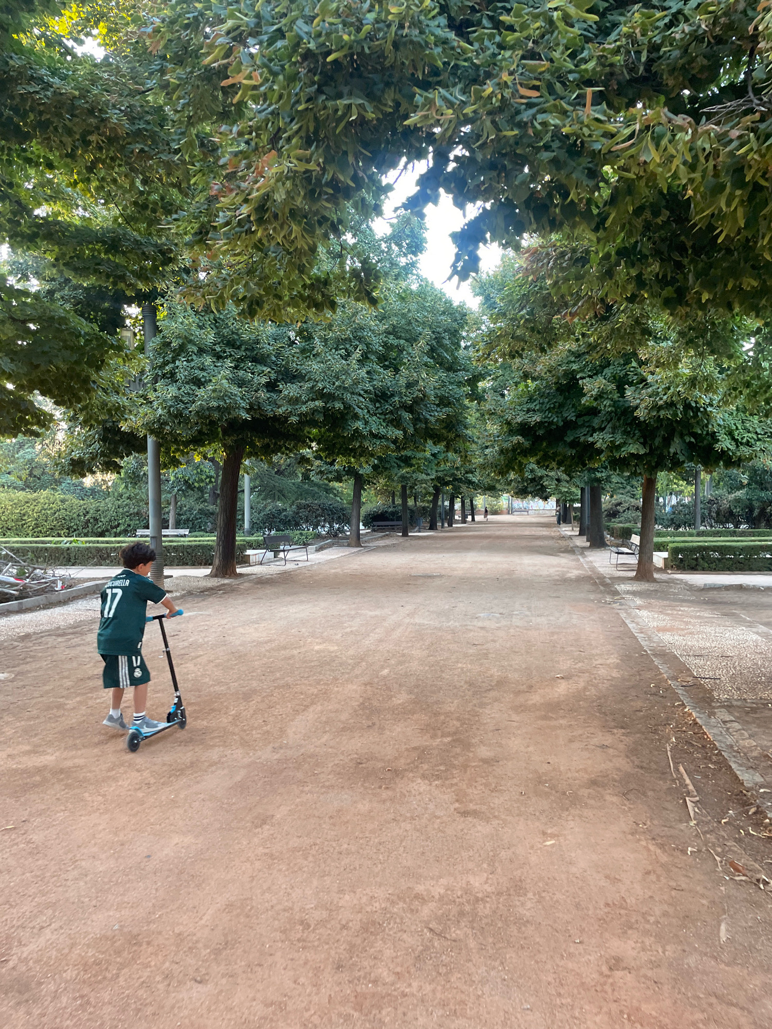 The image shows a young boy on a scooter in a park. The boy is wearing a green jersey with the number 17 on the back and dark shorts. He is positioned in the left foreground of the image, with his back to the camera, riding a blue and black scooter on a dirt path. The path leads into a tree-lined avenue with benches on either side. The trees are large and leafy, with sunlight filtering through the canopy. In the background, there are more trees and some buildings. The overall impression is one of a peaceful park setting. The image appears to be taken in the Generalife gardens in Granada, Spain, based on the distinctive architecture and landscaping.