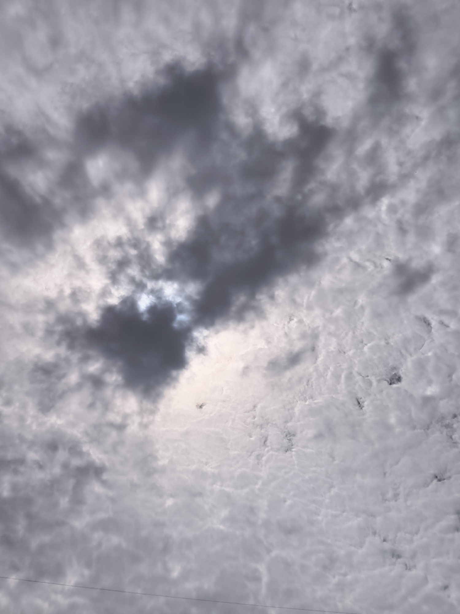 The image shows a cloudy sky. The clouds are a mix of dark grey and white, with some areas appearing brighter where the sun is peeking through. The overall impression is of an overcast or partly cloudy day. There are no people, subjects, or objects clearly visible in the sky. A thin, dark horizontal line, possibly a wire, is visible near the bottom of the frame. The image does not provide any specific visual cues to determine its location within the city of Krasnoyarsk, Russia.