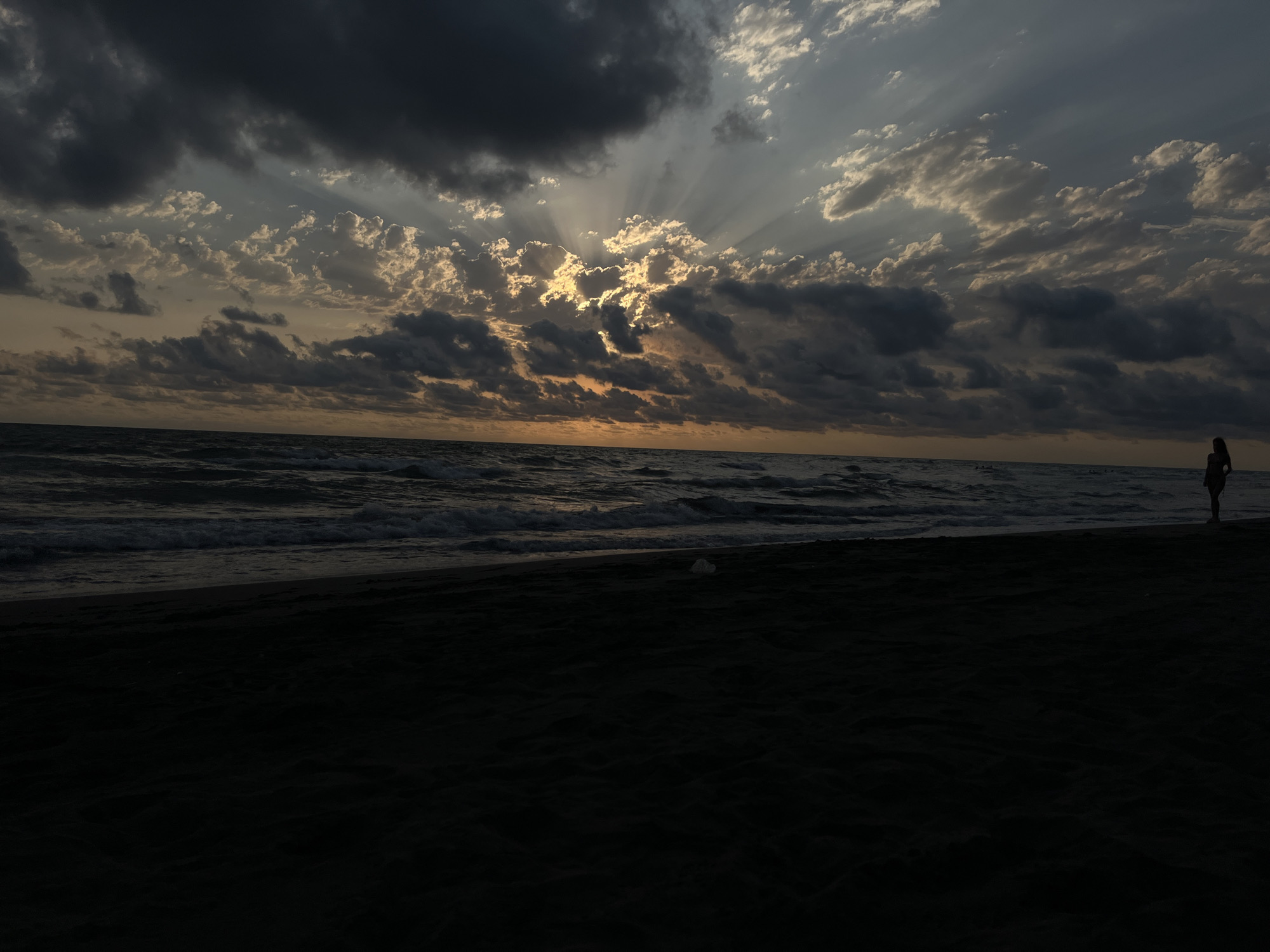 The image depicts a beach scene with a dark, sandy shore in the foreground and the sea in the background. The sky is dramatic, filled with a mix of dark, ominous clouds and lighter, scattered clouds through which the sun's rays are beaming. The sun appears to be low on the horizon, suggesting either sunrise or sunset. A lone figure, appearing to be a person, is standing on the beach near the water's edge on the right side of the frame. The waves are visible breaking on the shore. The overall mood is serene and somewhat melancholic due to the lighting and cloud cover. The location is the beach in the settlement of Supsa, Georgia.