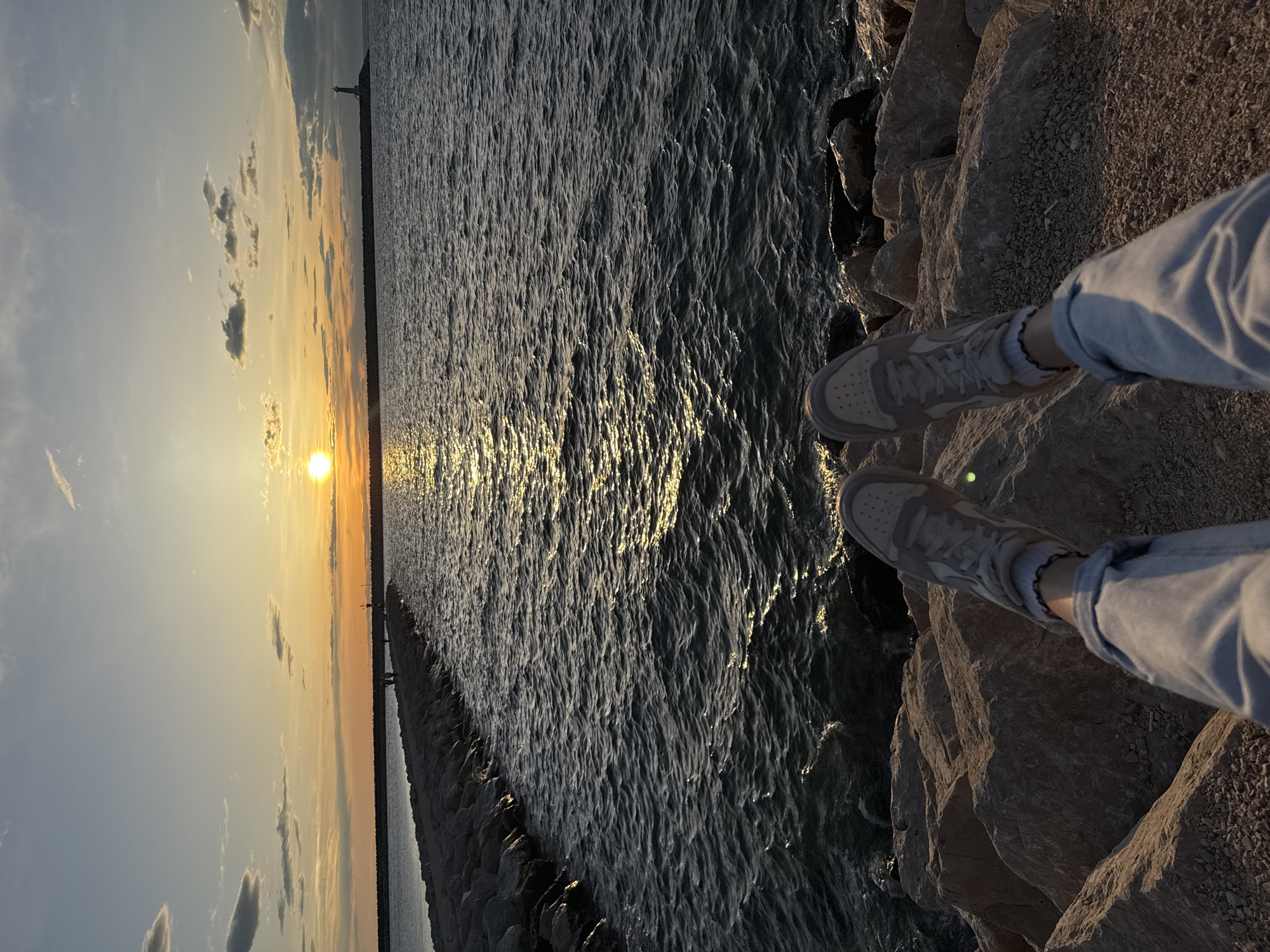 The image depicts a sunset scene on a rocky shoreline in Bar, Montenegro. The sun is setting over the water, creating a golden reflection on the surface. Two legs wearing light-colored sneakers and blue jeans are visible in the lower right corner, positioned on the rocks at the edge of the water. A stone pier extends into the water toward the sunset. The sky is a mix of light blue and orange hues, with some clouds scattered around the sun.