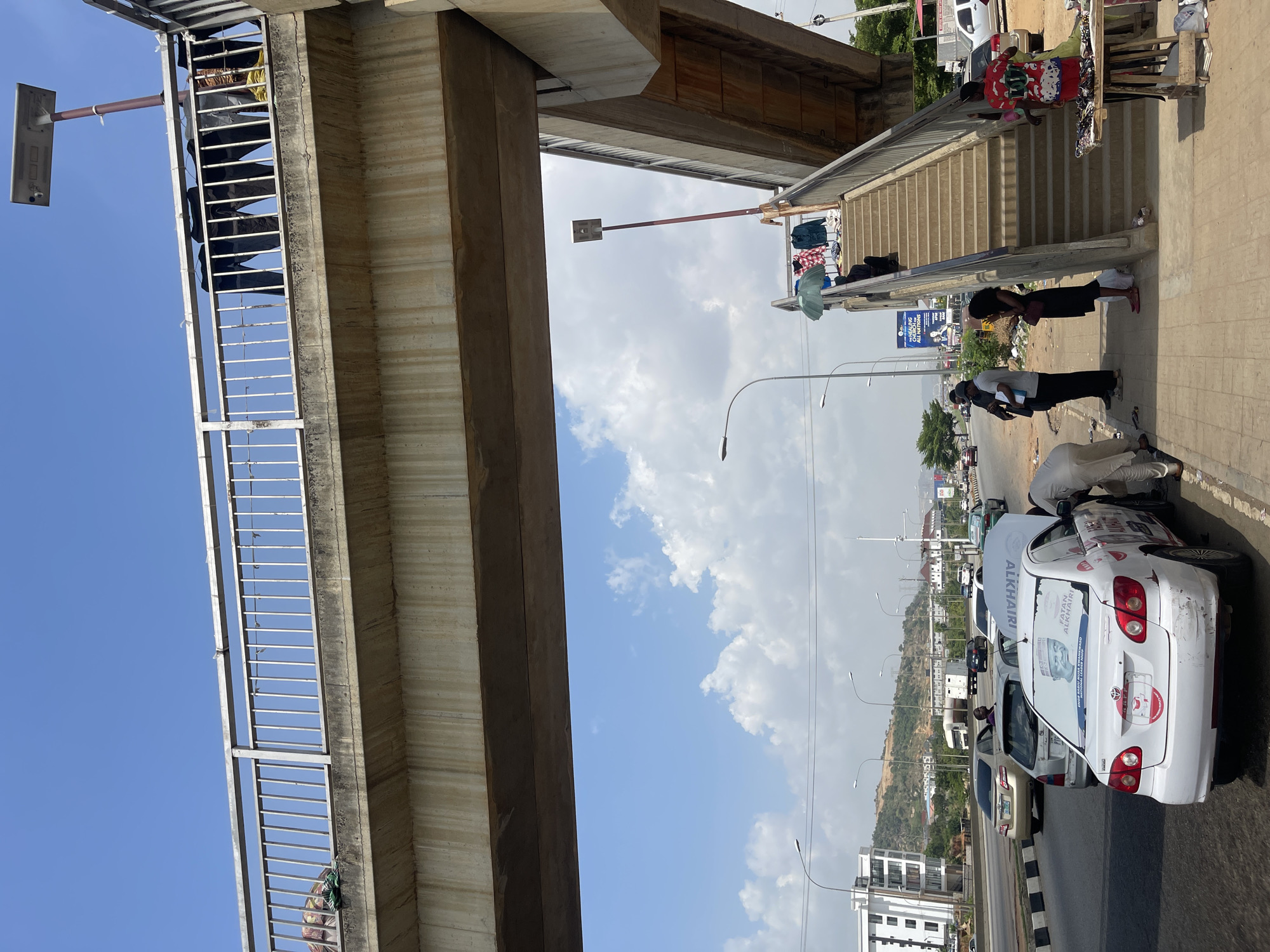 The image shows a pedestrian overpass in Gwarinpa, Nigeria. The overpass is made of concrete and steel, with stairs leading up to the walkway. People are walking on the sidewalk next to the overpass, and some are praying. Cars are parked on the side of the road, including a white car with political signage. In the background, there are buildings and hills. The sky is blue with some clouds. Clothes are hanging from the overpass.