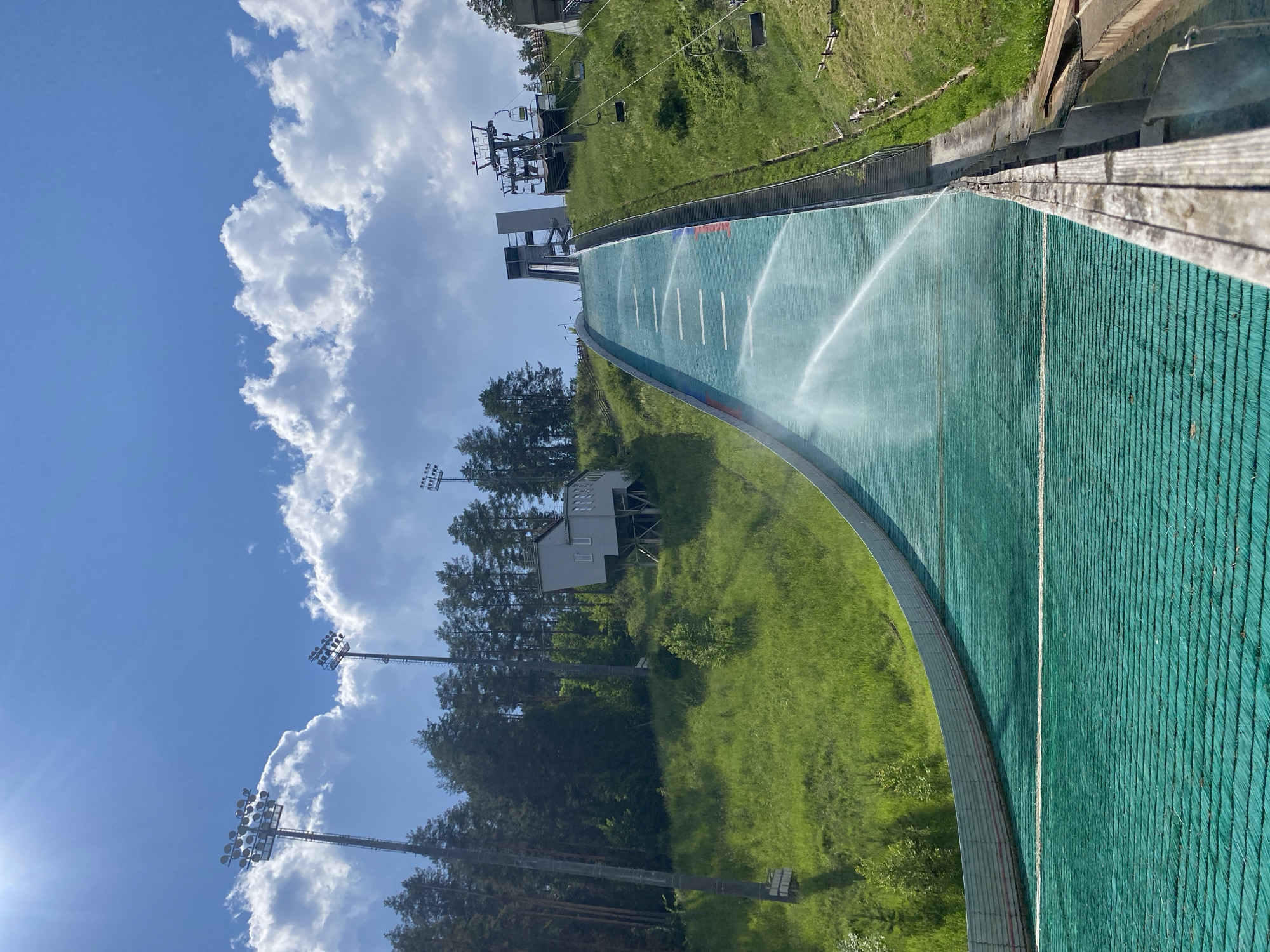 This image depicts a ski jumping hill located in the city of Perm, Russia. The hill is covered with green artificial turf, and water is being sprayed onto it from sprinklers. The ski jumping structure is tall and imposing, with a steep in-run and a large landing area. In the background, there are lush green trees and a bright blue sky with white clouds. There are also ski lifts visible on the side of the hill, suggesting this is a sports facility. The overall scene is one of a sunny day at an outdoor sports venue.