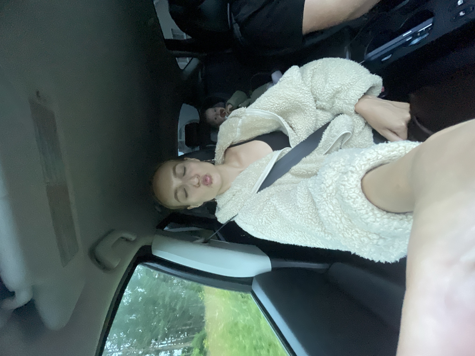 A selfie is taken from inside a car. A young woman with blonde hair is in the foreground, making a duck face. She is wearing a cream-colored fuzzy jacket over a black tank top and is buckled up with a seatbelt. Behind her, a baby sits in a car seat in the back, holding a pacifier. The car appears to be driving through a rural area with trees and greenery visible outside the window.