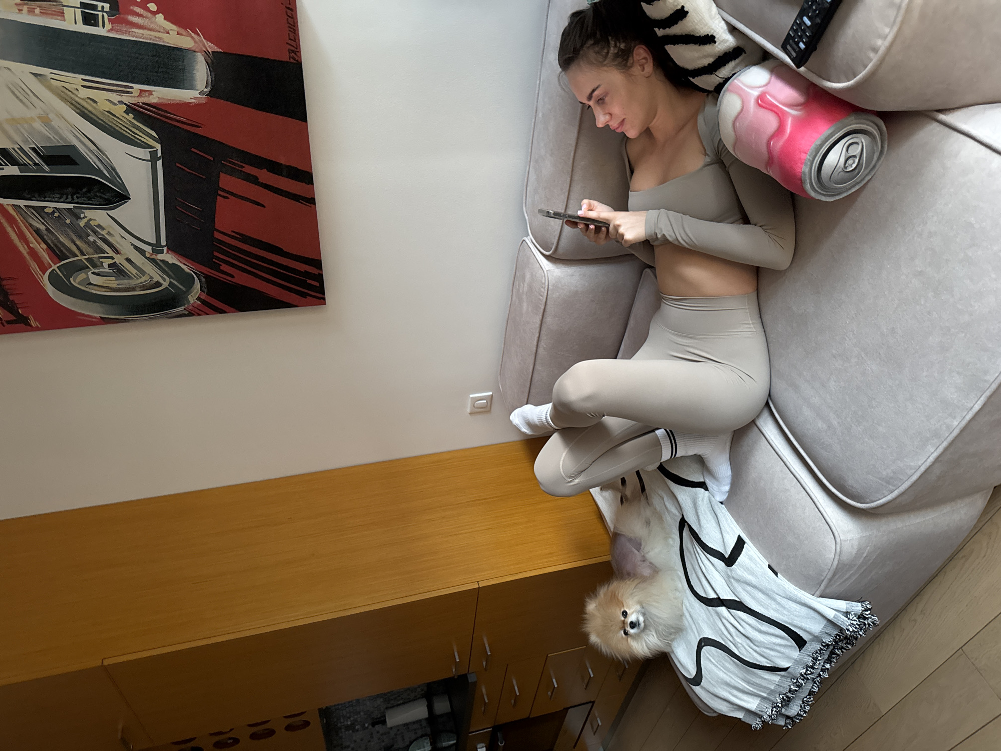 The image captures an indoor scene from a high, slightly tilted perspective, showing a relaxed moment in what appears to be a living room in Warsaw, Poland. A young woman with dark hair, wearing a light beige long-sleeved crop top and matching leggings with white and black striped socks, is lying on a plush, light-colored sofa. She is engrossed in her smartphone, holding it with both hands and looking down at the screen. Her expression is calm and focused.
Beneath her legs, nestled on a white blanket with a black abstract pattern, lies a small, fluffy, light brown Pomeranian dog, appearing very comfortable on its back with its legs slightly curled, looking towards the camera. Next to the woman's head on the sofa, there's a novelty pillow shaped like a pink and white soda can, and a dark TV remote control.
The background features a segment of a white wall, adorned with a large, vibrant piece of art. This artwork displays a stylized, industrial-looking machinery design in bold red, black, and white/grey, with the text "FALCUCCI" visible on its right edge, likely the artist's signature. Below the wall art, a long, low wooden cabinet or dresser with multiple compartments and silver handles extends across the lower portion of the frame. The floor is a light wood or laminate. The lighting suggests it is daytime, with soft, natural light illuminating the scene.