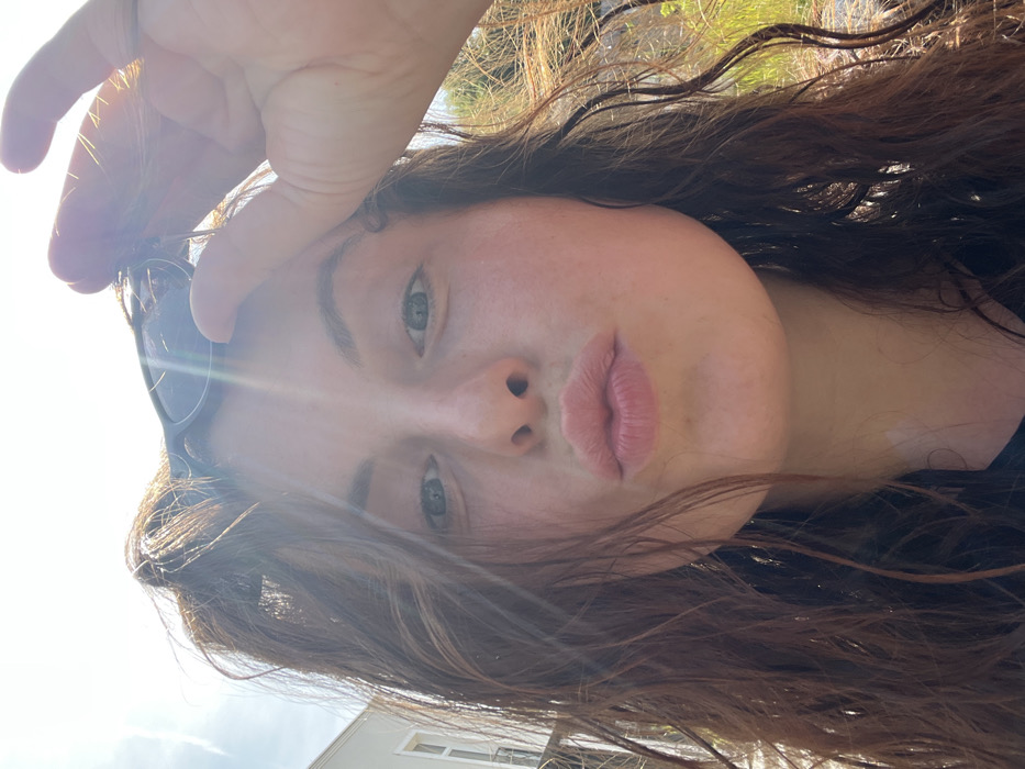 A close-up portrait, captured in Bad Sobernheim, Germany, features a single female subject with fair skin, blue eyes, and long, wavy brown hair. Her lips are pursed in a "kiss" gesture, and she looks directly at the camera. Her right hand is elevated, holding dark-framed sunglasses positioned atop her head. The scene is brightly lit by natural sunlight, causing visible lens flares. The background environment consists of a light-colored building with a visible window on the left, and green foliage against a clear blue sky on the right.