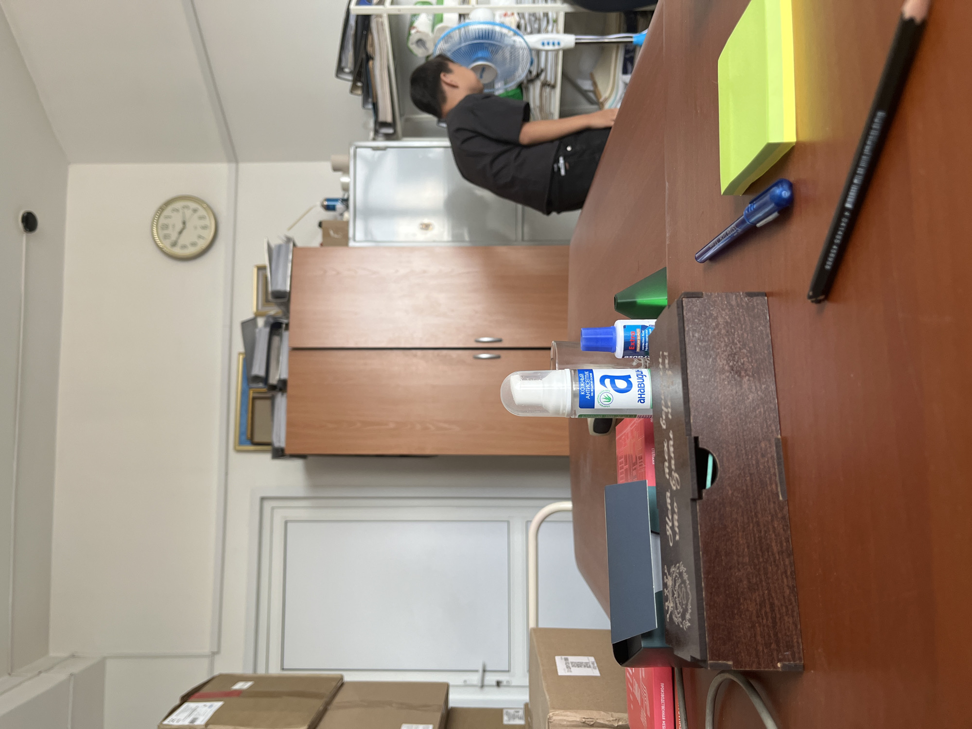 The image depicts an indoor scene, likely an office or storage area. In the foreground, a wooden desk is laden with various items. A dark wooden box with intricate carvings sits prominently, alongside a stack of red and white papers, and a blue bottle of correction fluid. A blue pen and a stack of bright yellow sticky notes are also visible on the desk. Further back, a person wearing a black t-shirt and dark pants is partially visible, appearing to interact with a blue and white oscillating fan. To the left, stacked cardboard boxes are piled high, and a white door with a glass panel is visible. In the background, a large wooden cabinet stands against a wall, and a round clock with Roman numerals is mounted high on the wall. Shelving units filled with files and binders are also present. The environment appears to be lit by artificial light, casting subtle shadows.