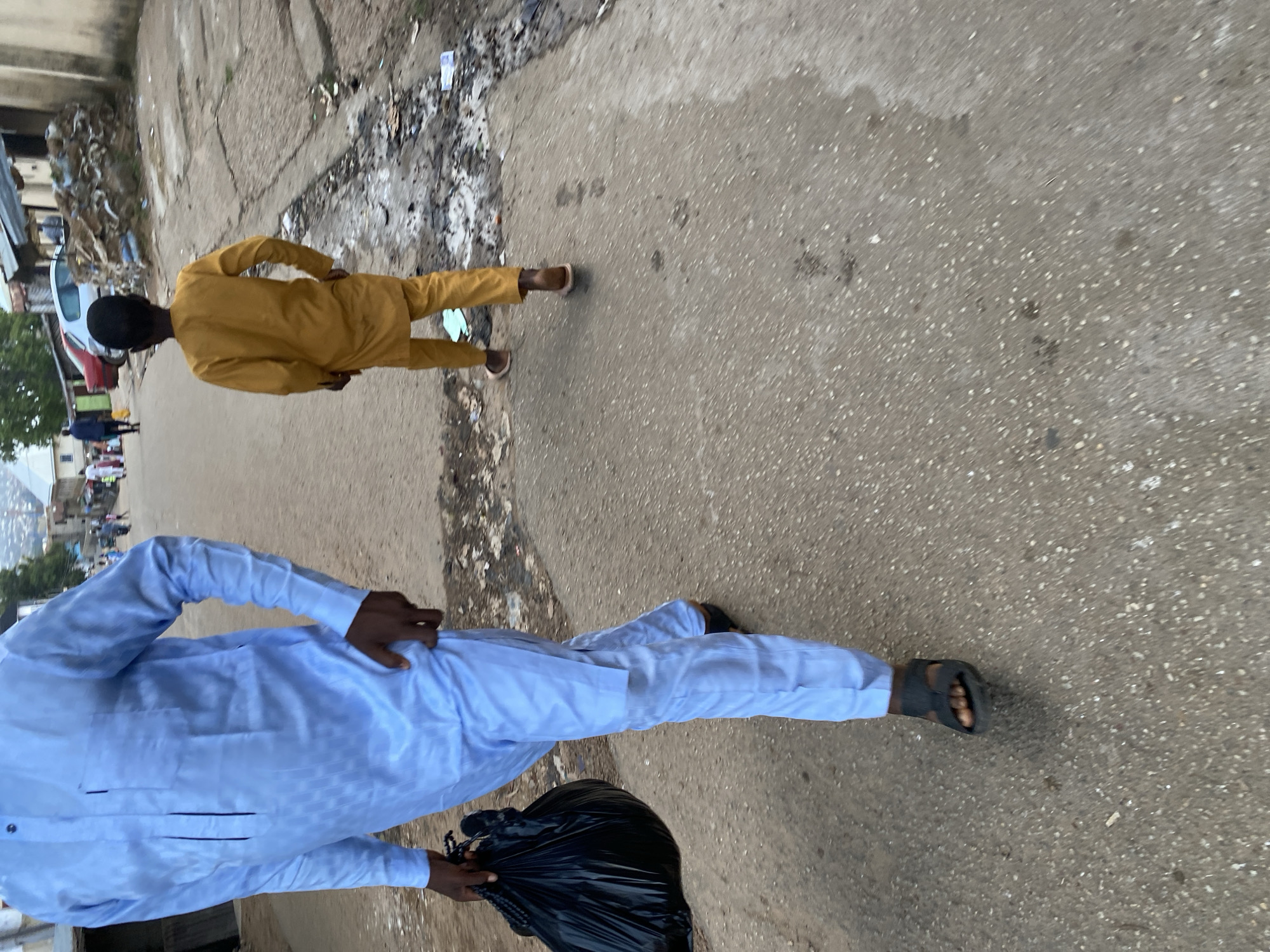 In Jos, Nigeria, an adult in a light blue, long-sleeved outfit and dark sandals walks on a paved road, carrying a black plastic bag in their left hand. A child in a mustard-colored outfit walks ahead on the same road. The environment appears to be an urban area with buildings and debris visible on the side of the road. The lighting suggests it is daytime, possibly overcast or late afternoon.