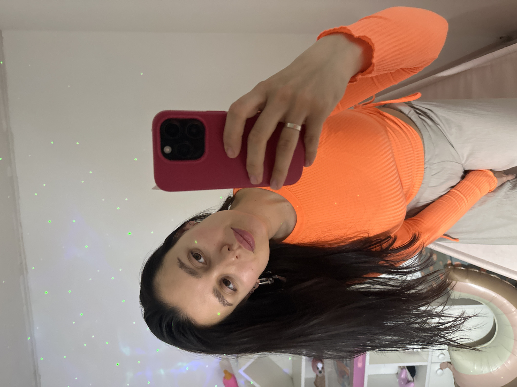 The image, taken in Chaikovsky, Russia, features a young woman capturing a mirror selfie indoors. The photograph is oriented vertically, showing the woman's head tilted towards the lower left. She has long, dark hair and gazes directly at the camera with a neutral expression. She is dressed in a vibrant orange, ribbed, long-sleeved crop top paired with light-colored (possibly beige or light grey) pants. A simple silver-toned ring adorns her left ring finger. She holds a smartphone encased in a bright pink/red cover, clearly displaying its triple camera array, in her left hand.
The background behind her is a plain white wall, illuminated by numerous scattered green light dots, suggestive of a laser star projector. On the right side of the frame, parts of a large, inflatable or plastic ring-shaped object, striped in light brown and pinkish hues, are visible. Beneath this object, shelves or cubbies containing a pink item can be faintly discerned. The setting appears to be an interior space, possibly a bedroom, with soft ambient lighting. No text is visible in the image.