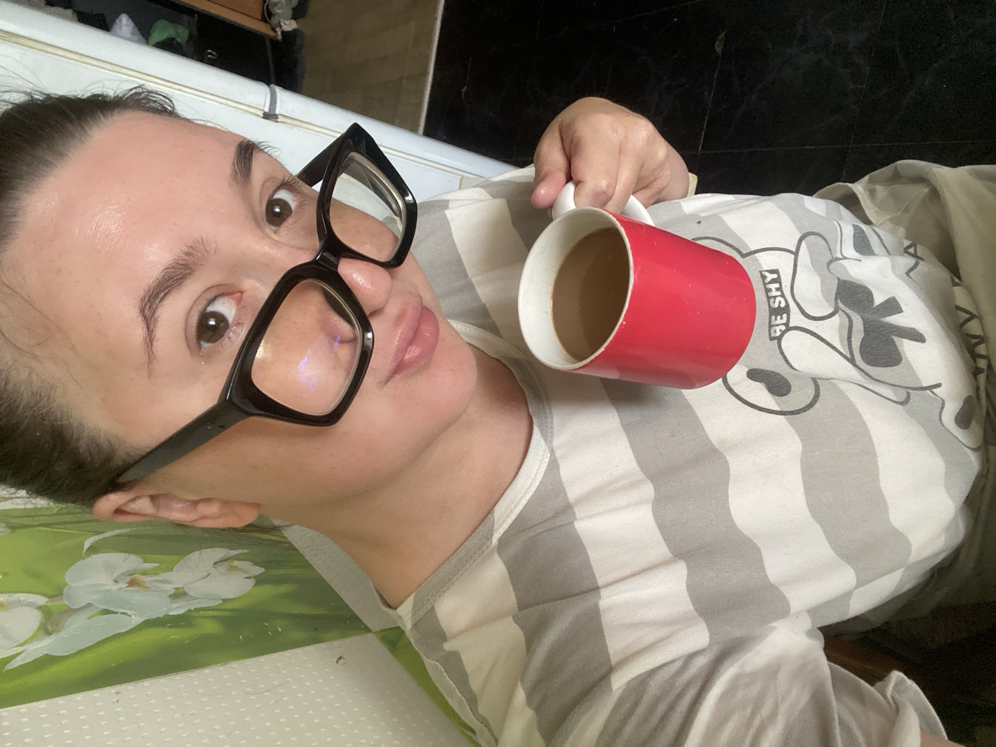 This media file presents a close-up, selfie-style portrait of a person, likely a woman, captured from an overhead perspective while she is lying down or reclining. The image is rotated, with her head positioned towards the left. She has dark hair, pulled back, and is wearing large black-framed glasses. Her eyes are open and gaze directly at the camera.
She holds a bright red mug, filled with a light brown beverage (likely coffee or tea), in her right hand. She is dressed in a casual t-shirt with alternating grey and white stripes. On the t-shirt, a graphic, possibly a cartoon character, is partially visible, accompanied by the text "BE SHY". Her expression is relaxed, featuring a slight pout, suggesting a casual or playful demeanor.
The background indicates an indoor setting. To the left of her head and body, a light green surface with a white floral pattern is visible, potentially a bedspread, pillow, or wall decoration. To her right and extending behind her, a dark, possibly tiled, floor is present, along with some indistinct dark and white elements that could be furniture or parts of a room. The scene depicts a casual, domestic moment. No specific time of day or weather conditions can be determined from the image. This scene is located in Chaikovsky, Russia.