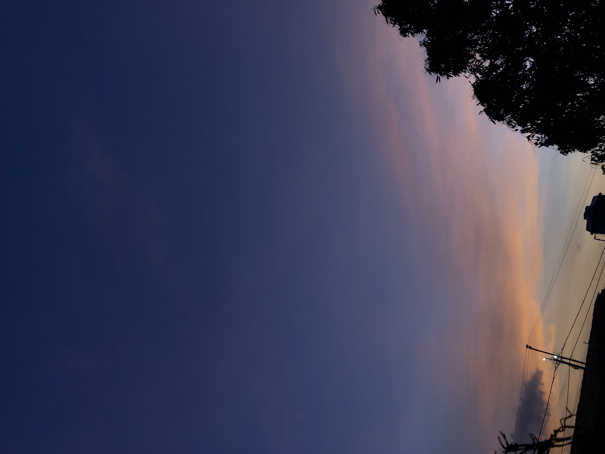 The image shows a dusky sky, transitioning from deep blue to orange and pink hues towards the horizon. A silhouette of a tree occupies the upper right corner. Utility poles and wires are visible along the lower right edge, with a building outline. The location is Altagracia, Venezuela. No people are visible. The scene depicts a moment at dusk.