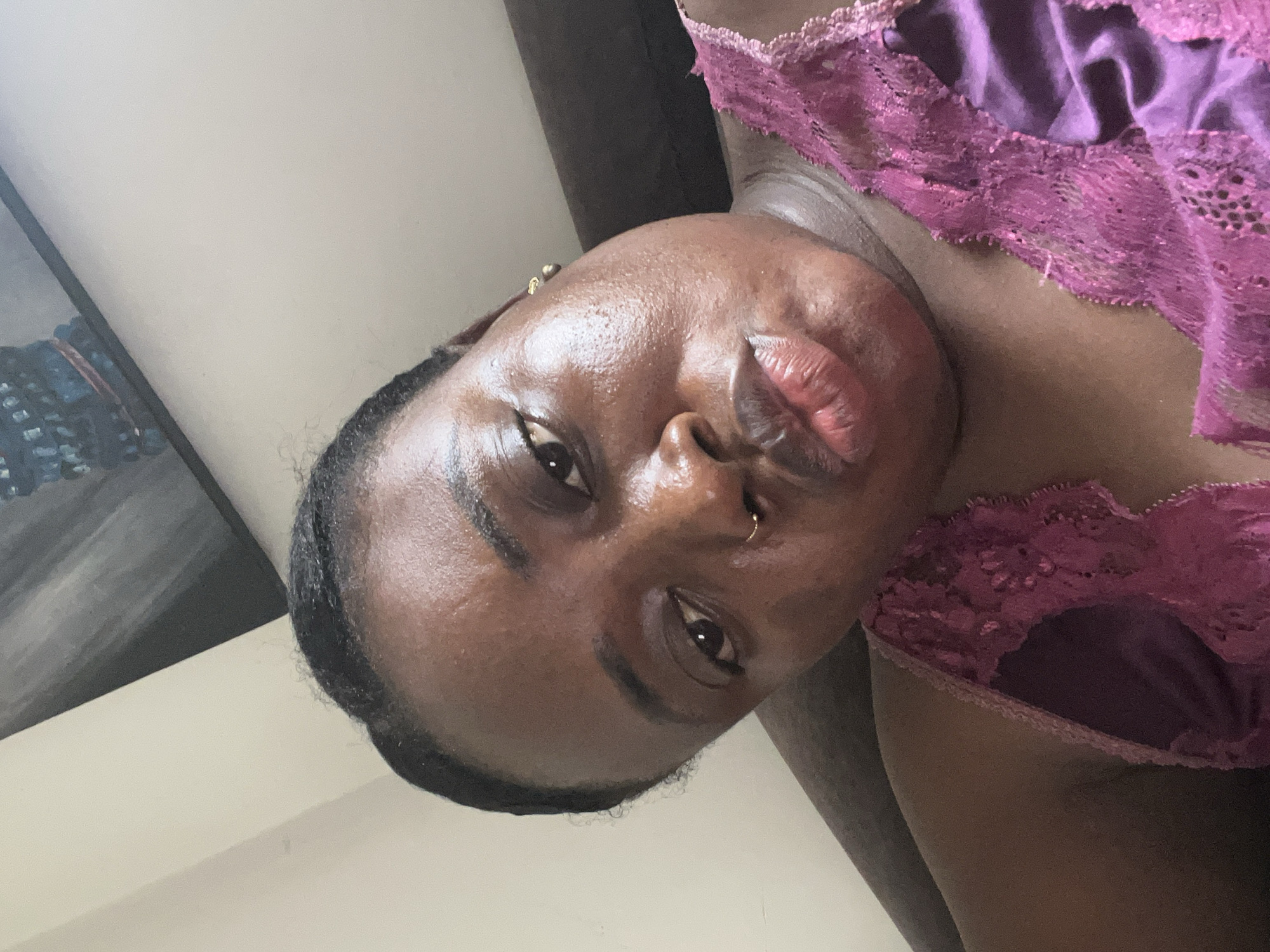 A close-up, rotated selfie shows a dark-skinned person with a nose ring, wearing a maroon lace-trimmed top. The background includes a light-colored wall and a dark, indistinct object in the upper left corner. The location of this image within Abuja, Nigeria, cannot be determined from the provided visual information.