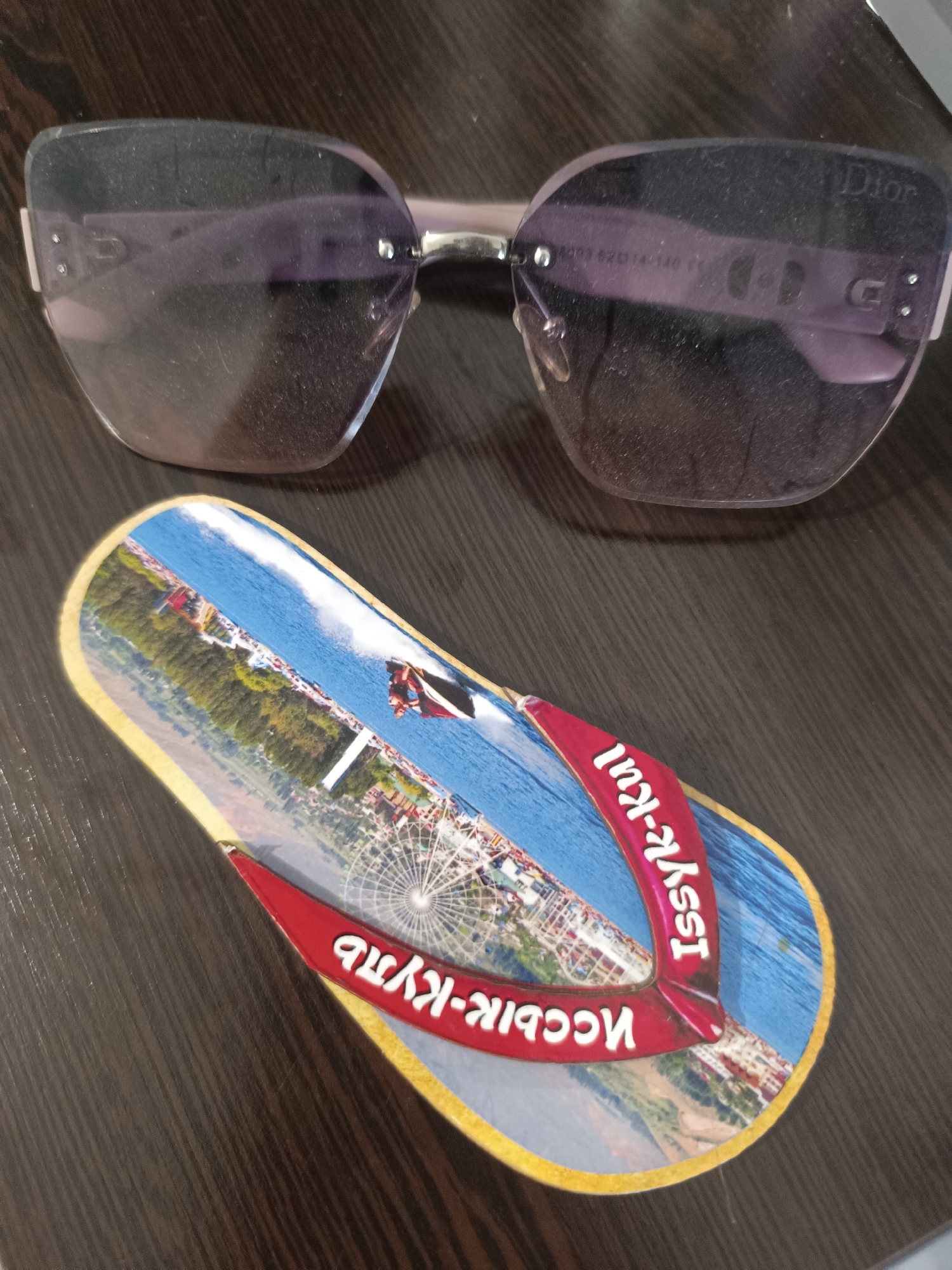 A pair of light purple Dior sunglasses with silver accents rests on a dark wooden surface. Next to the sunglasses is a souvenir flip-flop shaped object adorned with a panoramic image of a body of water, a Ferris wheel, and people engaged in water sports. Text in an unfamiliar script is visible on the flip-flop, and the sunglasses have text engraved on the lens. The scene is indoors and appears to be a flat lay composition.