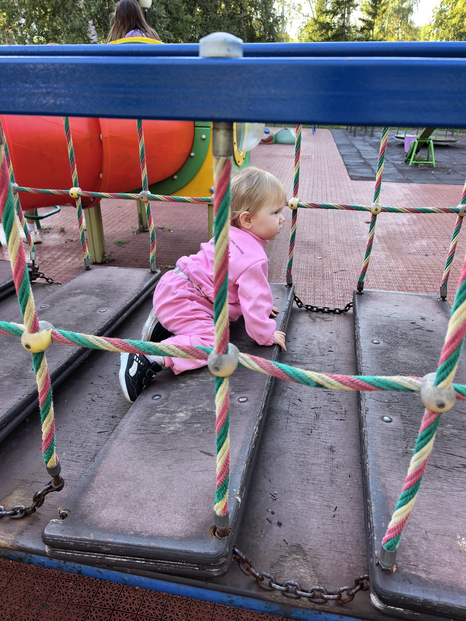 In Sterlitamak, Russia, a young child wearing a pink outfit and black and white sneakers crawls on a wooden platform on a playground. The child is positioned on the right side of the image, facing towards the right. In the background, a large red and yellow play structure is visible, along with trees and other playground equipment. The foreground is dominated by a blue railing and colorful ropes with yellow connectors, part of the playground's climbing structure. The ground beneath the platform is a mix of dark wood and red patterned material. The scene appears to be outdoors during daylight.