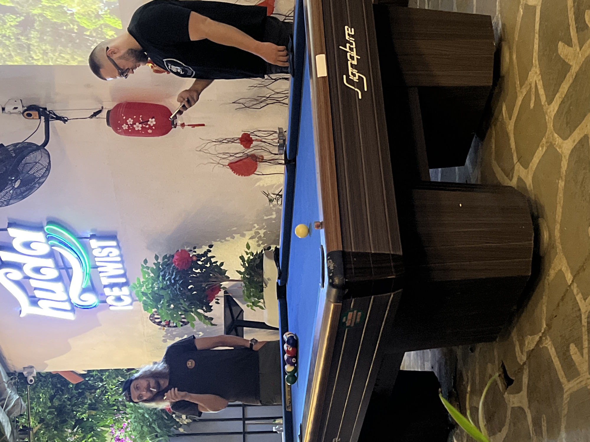 An indoor scene in Đà Nẵng, Vietnam, possibly a bar or entertainment establishment featuring a "huda ICE TWIST" neon sign. A pool table dominates the right side of the image. On the left, a man with long hair and a beard stands near the pool table, holding a cigarette. Another man stands at the opposite end of the table, looking at a phone or device. Decorations include paper lanterns and floral arrangements. The floor appears to be paved with patterned stones.