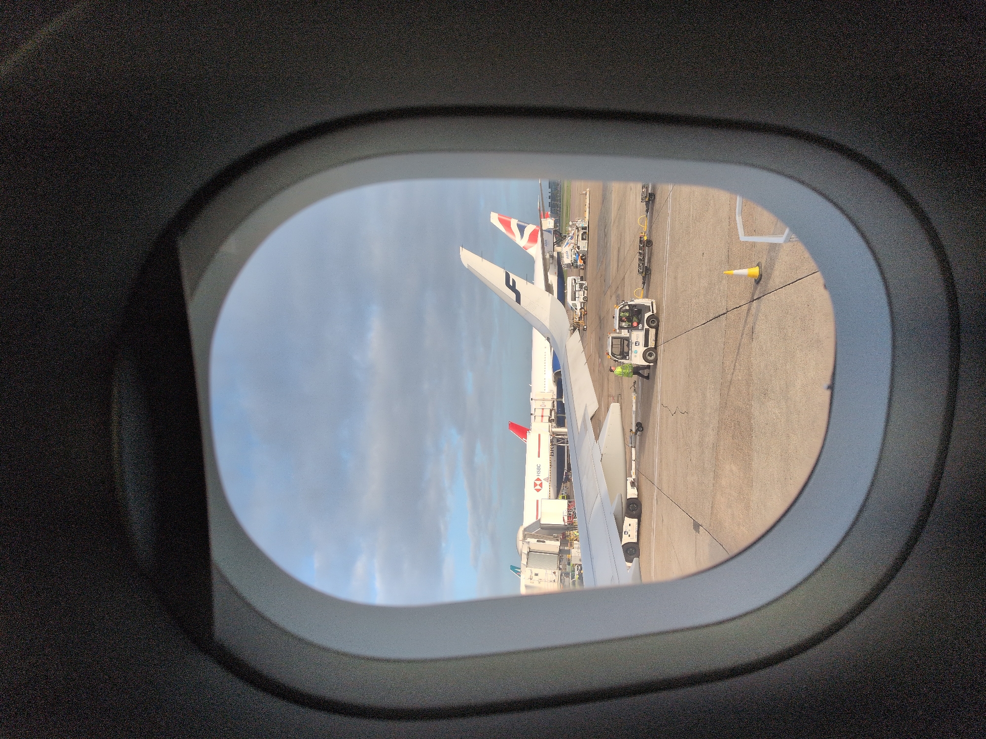 View through an airplane window. The wing of the airplane is visible in the foreground. In the background, other airplanes are at their gates. One is a British Airways plane with a Union Jack on the tail. Another plane is painted white with HSBC in red letters. Ground crew and ground vehicles are visible. The location is likely Heathrow Airport in London, United Kingdom.