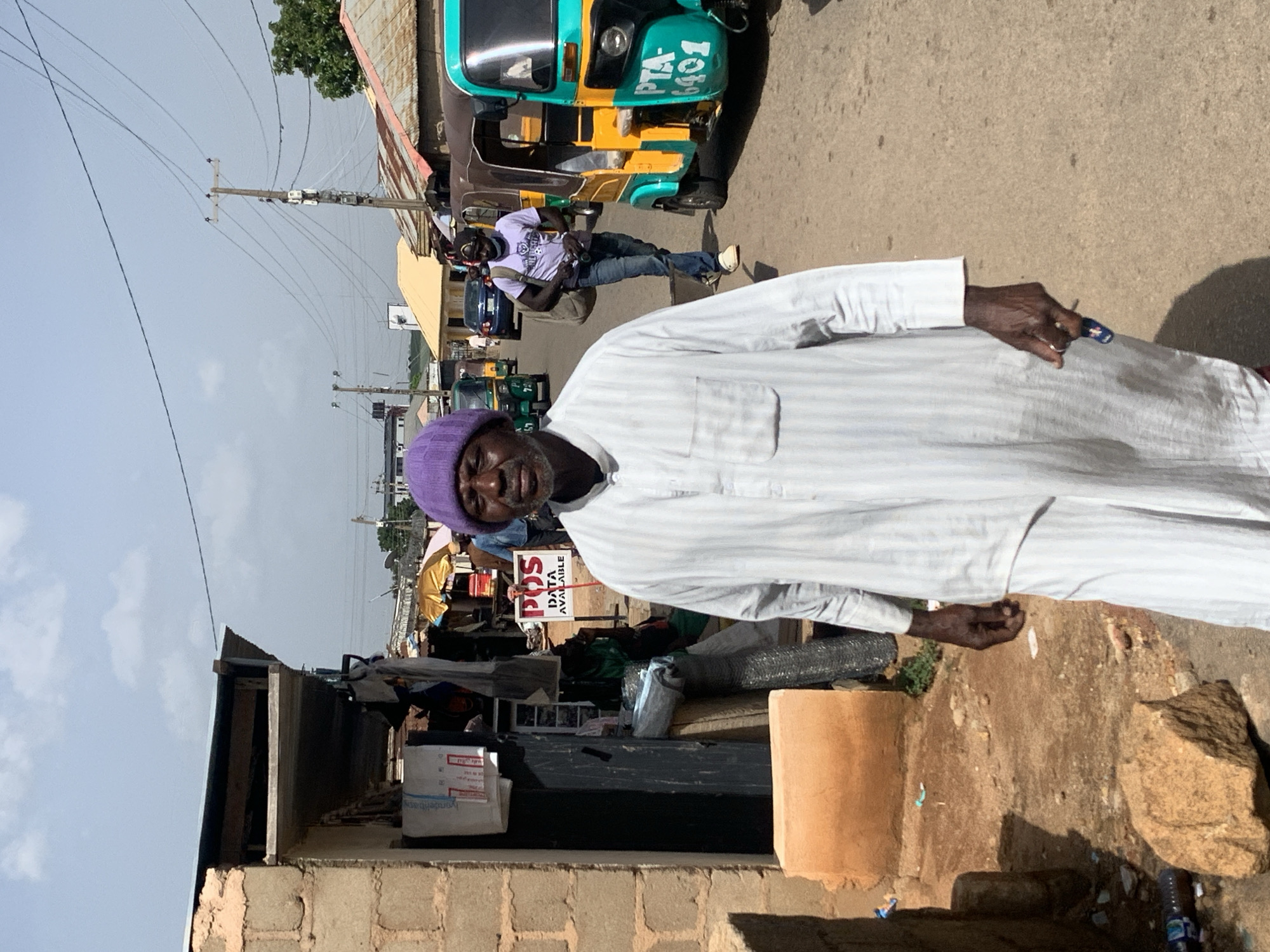 The image depicts a street scene in Jos, Nigeria. The primary subject is an elderly man wearing a white striped robe and a purple knit cap. He is looking towards the camera with a neutral expression. His right hand is visible, with a dark blue and white ring on his index finger.
Behind the man, the scene unfolds into a bustling street. A colorful auto-rickshaw, predominantly yellow and teal with the license plate "PTA 401" visible, is parked on the right side of the road. Another auto-rickshaw is further down the street. A man in a white t-shirt and jeans is standing near the yellow and teal auto-rickshaw, facing away from the camera, with a bag slung over his shoulder.
Various stalls and structures line the street, suggesting a market or commercial area. A sign with "POS DATA AVAILABLE" printed in red and white text is prominently displayed. There are electrical wires and poles overhead, indicating urban infrastructure. The ground is paved with asphalt, and there are some piles of debris and building materials visible, particularly on the right side of the frame, including large rocks and cylindrical objects.
The lighting suggests it is daytime, likely sunny, with distinct shadows cast by the objects and structures. The sky is overcast with some patches of blue. The overall impression is of a busy, everyday scene in a Nigerian town.