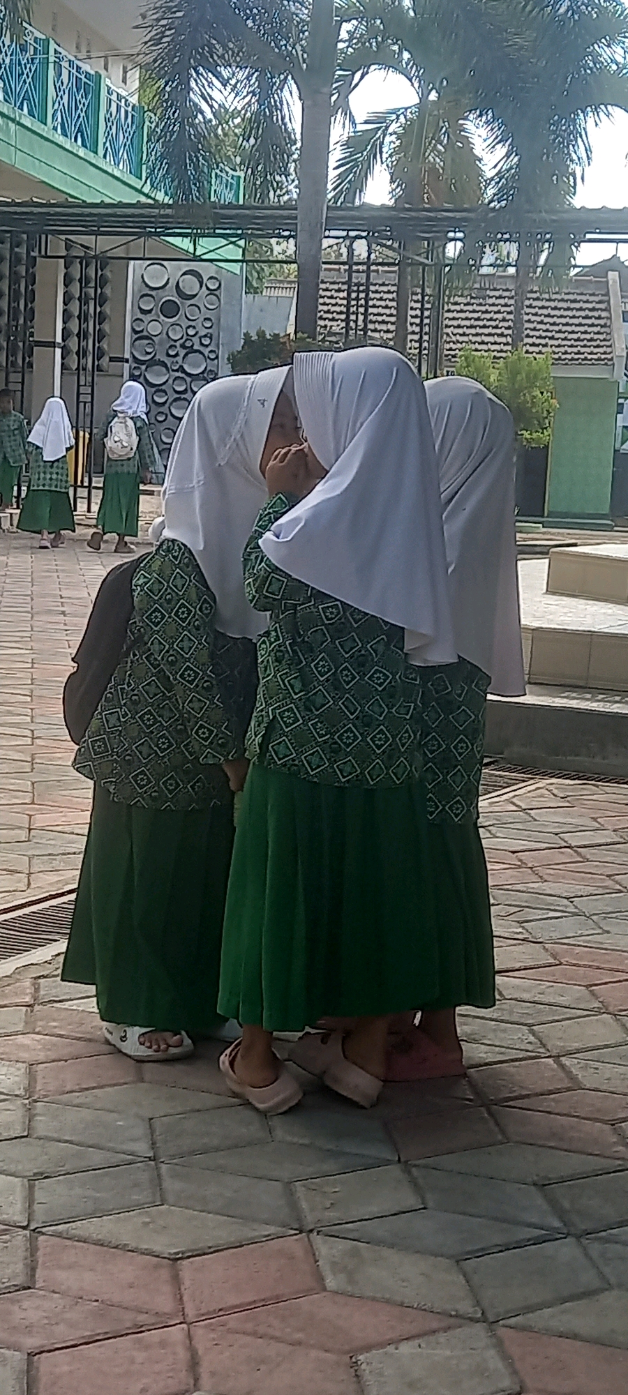 The image shows a group of young girls in school uniforms standing on a paved walkway in front of a building. The girls are wearing white hijabs and green dresses with a patterned design. The building appears to be a school with a modern facade, featuring a green and white color scheme and decorative metal railings. Palm trees are visible in the background. The ground is paved with interlocking hexagonal and rectangular tiles in shades of grey, pink, and beige. The lighting suggests it is daytime, likely outdoors. The girls in the foreground are clustered together, with two of them facing each other and one appearing to be talking or whispering. Other girls are visible further in the background, also in uniform. Based on the context of similar imagery, this scene is likely located at a school in Pontang, Indonesia.
