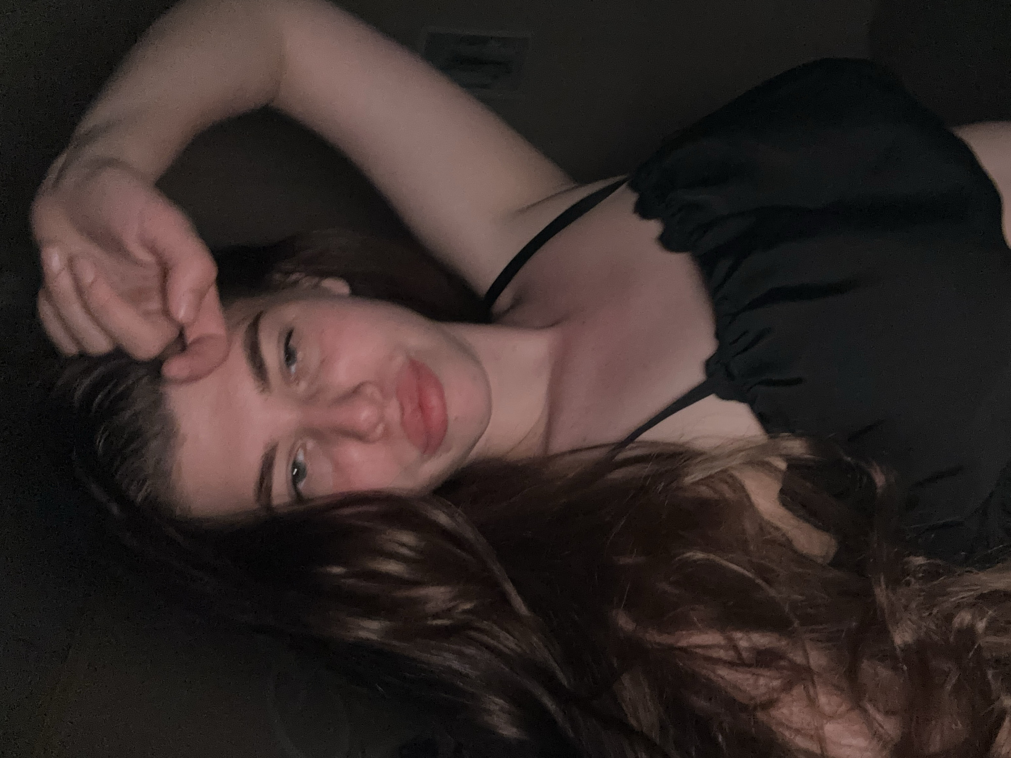 A woman with long brown hair is lying on her side, facing the camera. She is wearing a black top with thin straps. Her left arm is raised, with her hand near her forehead. The background is dark. The location is not discernible from the image.