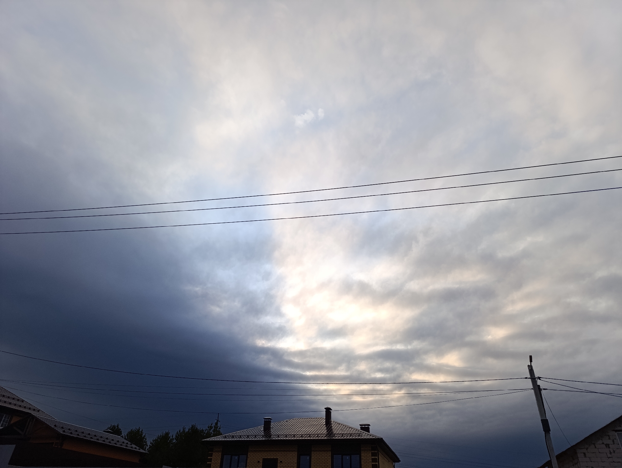 The image shows a cloudy sky over a residential area. In the foreground, a brick house with a brown tile roof is visible. There are two chimneys on the roof. Power lines run horizontally across the frame. A utility pole stands to the right of the house. Trees are visible in the middle distance. The sky is filled with dark and light gray clouds, with a patch of brighter, lighter sky towards the center. The location is Chaikovsky, Russia.