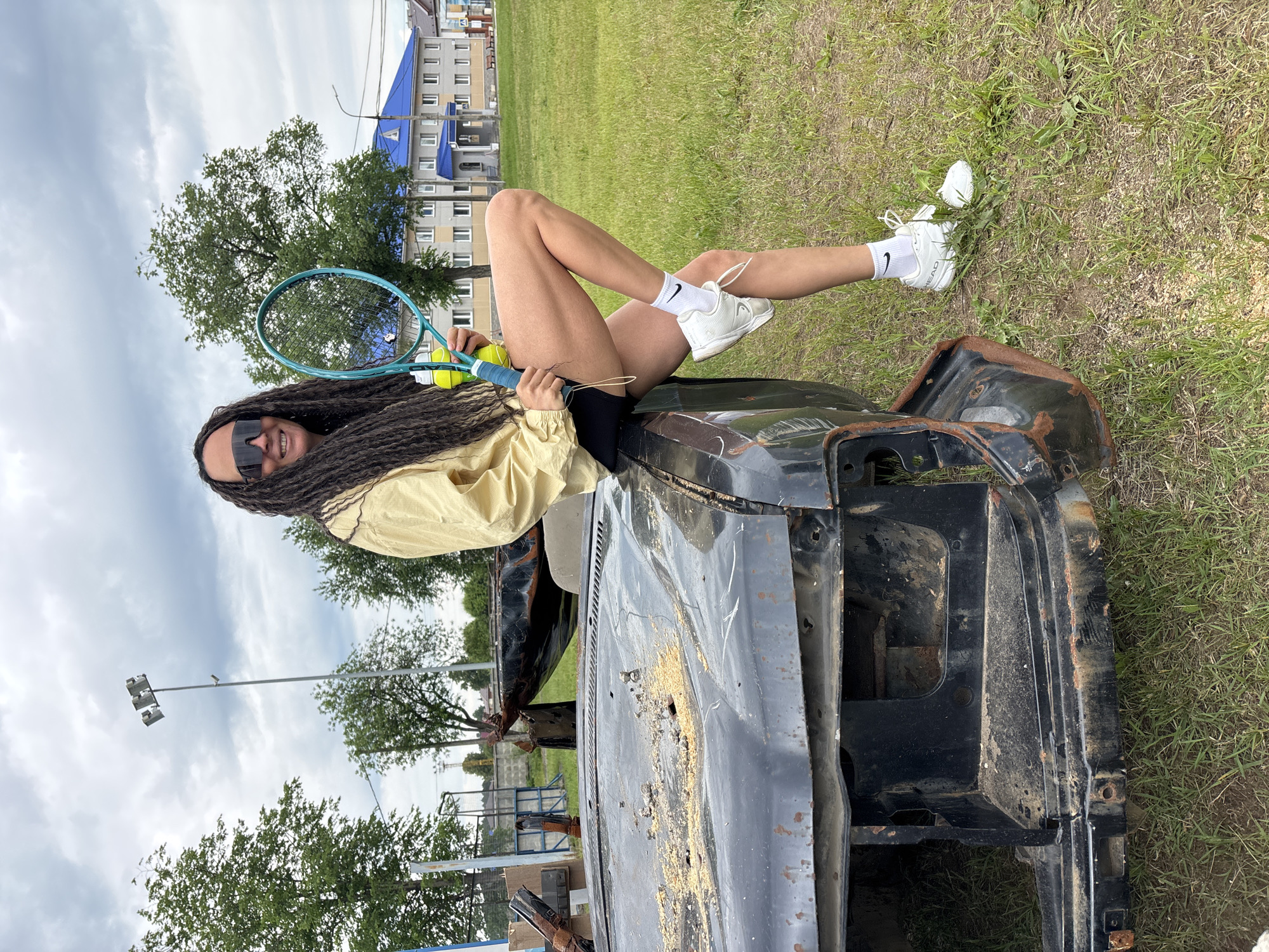An outdoor, eye-level shot depicts a person with long dreadlocks posing on a derelict vehicle. The person, wearing sunglasses, a light beige jacket, black shorts, white Nike socks, and white sneakers (with "HEAD" visible on one shoe), is seated on the rusted and damaged front section of a dark-colored car, holding a light blue tennis racket and two yellow tennis balls. Their legs are bent, with one foot touching the grassy ground beside the vehicle.
The car's front end is heavily weathered, with rust and what appears to be sand or dirt covering parts of the hood and chassis. The engine bay area is exposed.
The background features a large grassy field with scattered trees. In the distance, several multi-story buildings are visible, one distinctively topped with a bright blue roof. A tall light pole with floodlights stands in the mid-ground, suggesting a nearby sports facility or public area. The sky is overcast with moderate cloud cover, indicating daytime. The overall setting appears to be a somewhat neglected area, possibly an urban or suburban fringe in Chaikovsky, Russia. The person maintains a relaxed and confident posture while looking towards the camera.