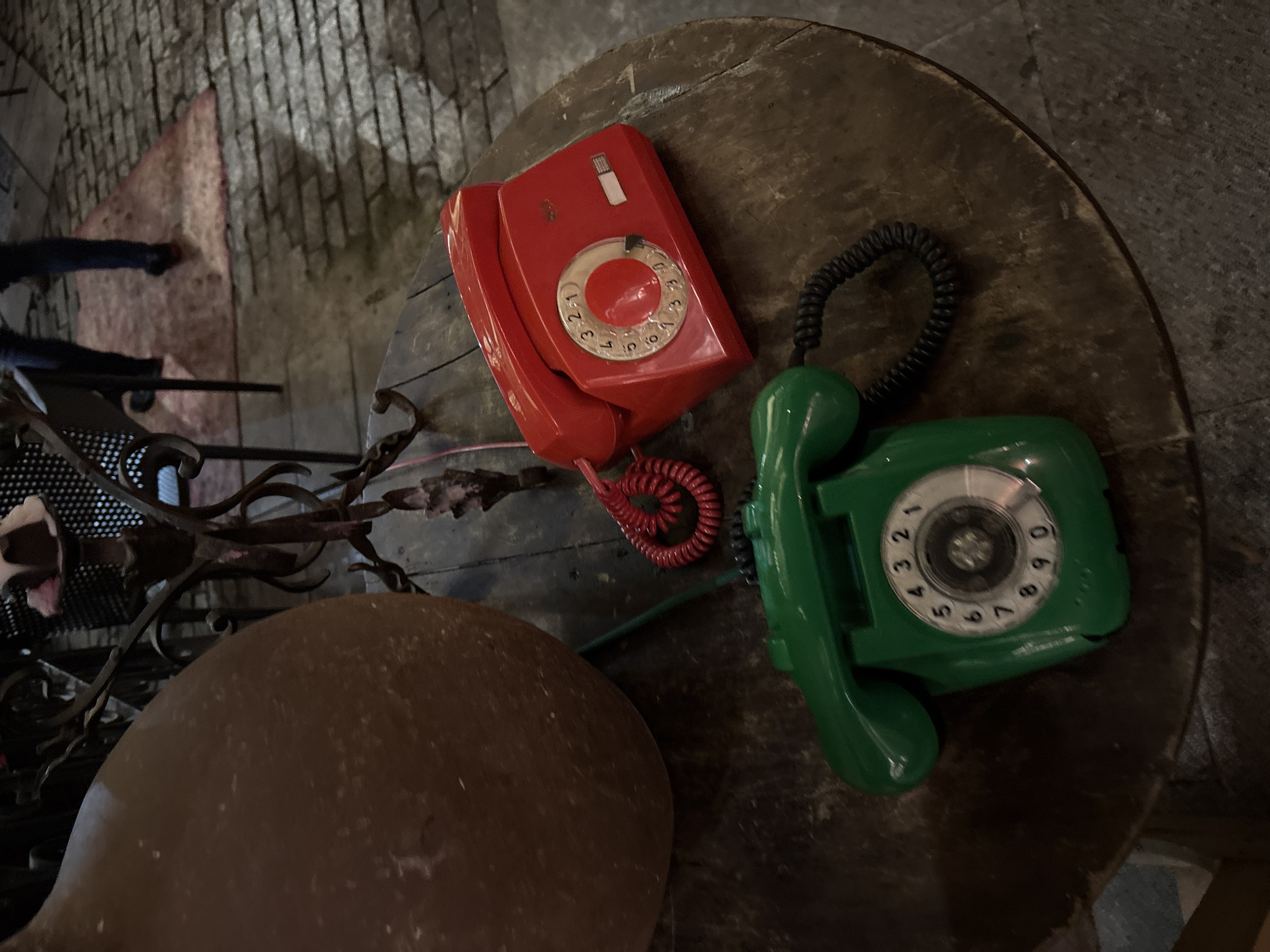 Two rotary telephones, one red and one green, are positioned on a round wooden table. The red telephone is on the right side of the table, while the green telephone is on the left. Both telephones have their cords connected. In the background, a person's legs are visible, wearing dark pants and shoes, standing on a cobblestone surface with a patterned rug. A large brown ceramic pot is also visible on the left side of the table, behind the green telephone. The scene appears to be outdoors, possibly in a public area given the cobblestone pavement and general ambiance. The lighting suggests it may be evening or indoors with dim lighting.